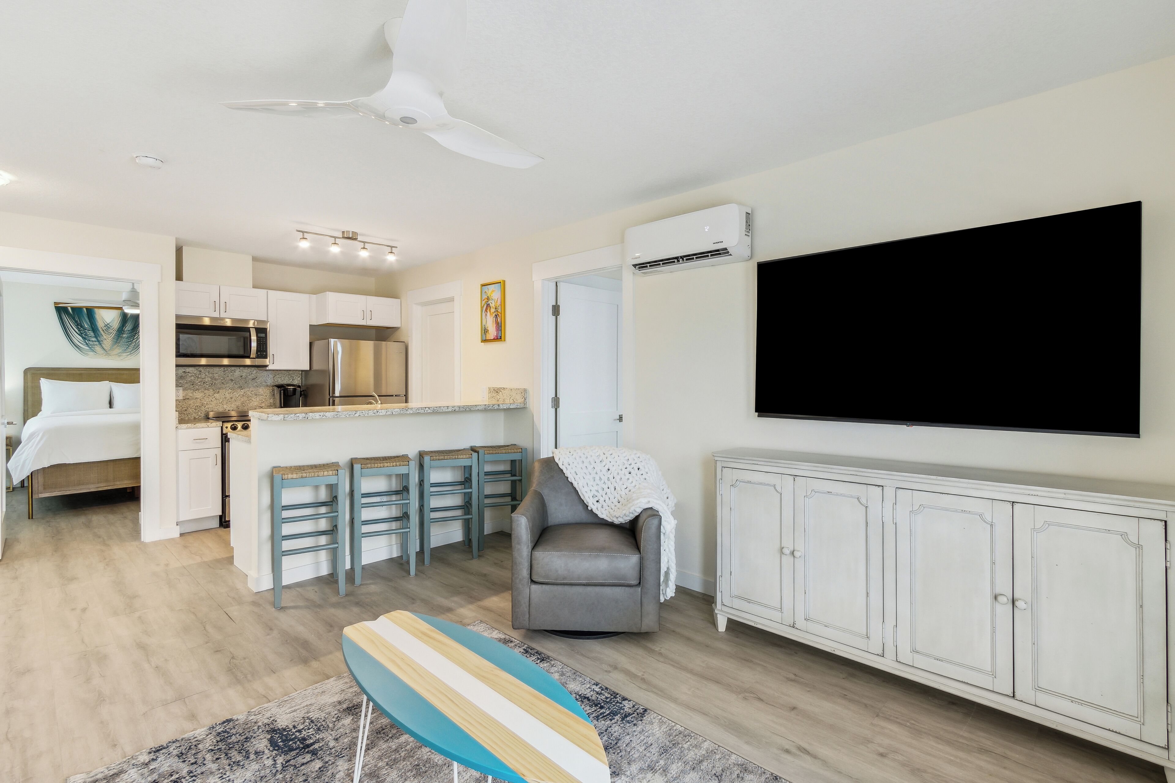 A modern kitchen with a microwave, stove, and white cabinets. There is a dining area with a table and chairs, and a gray armchair. A flat-screen TV is mounted on the wall.