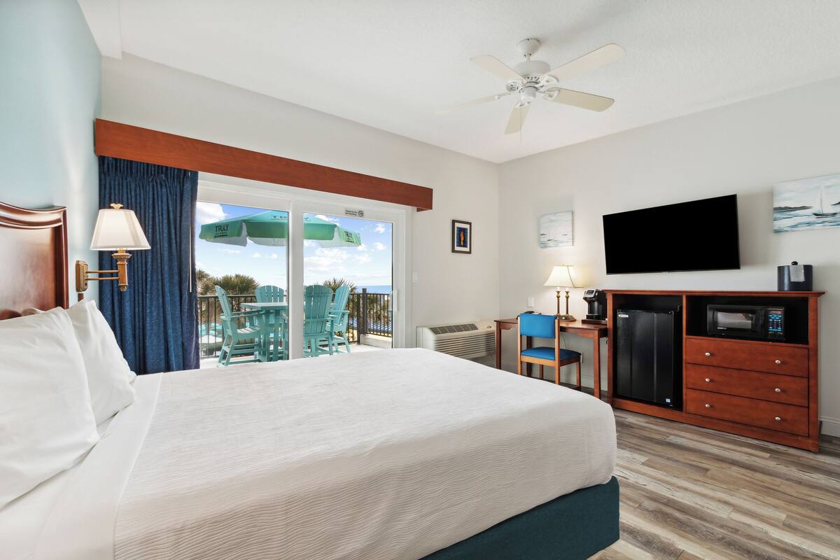 A hotel room with a large bed, a TV, a minibar, and a balcony with chairs and a table.