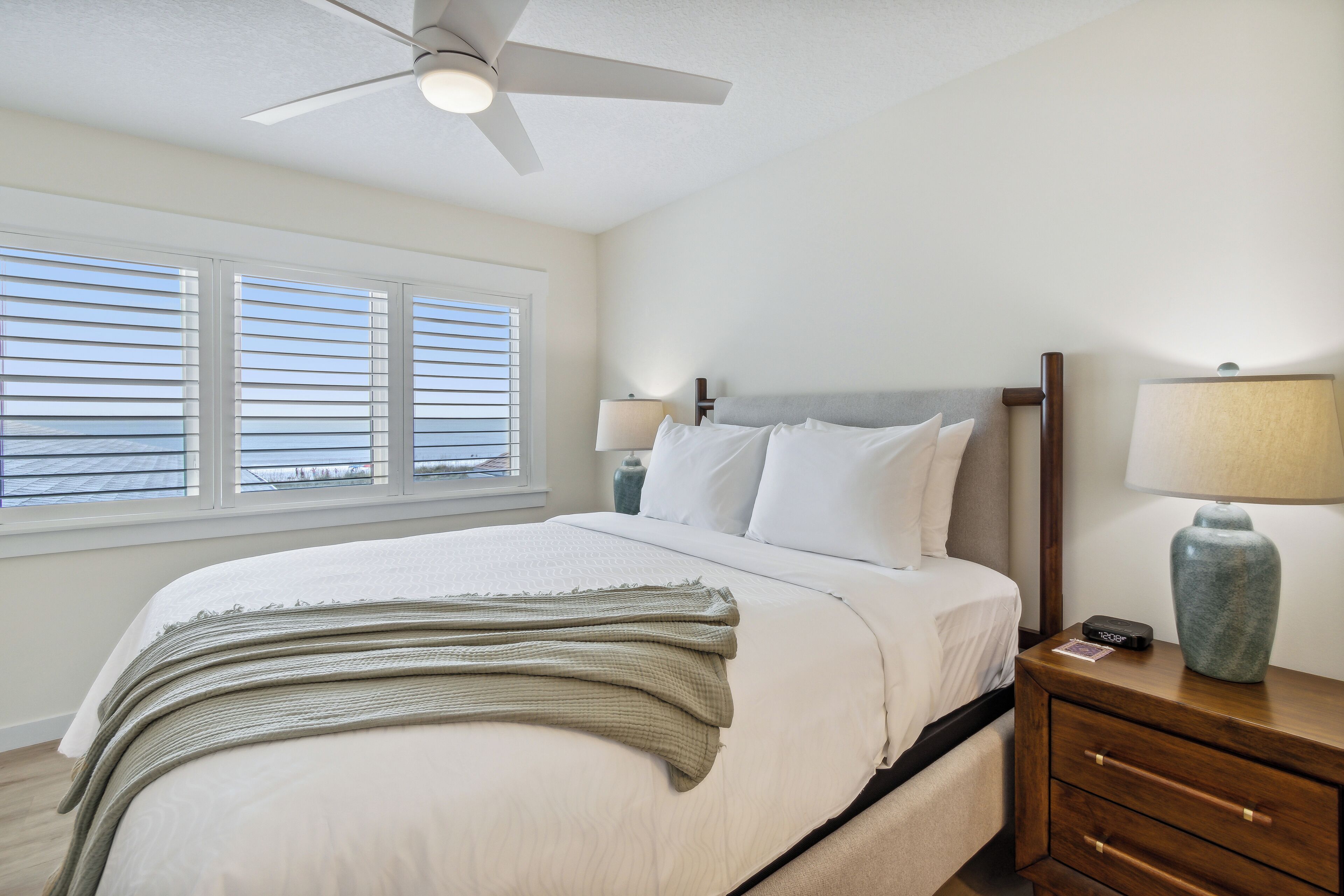 A neatly made bed with white linens and a green throw blanket, a bedside table with a lamp, and a ceiling fan.