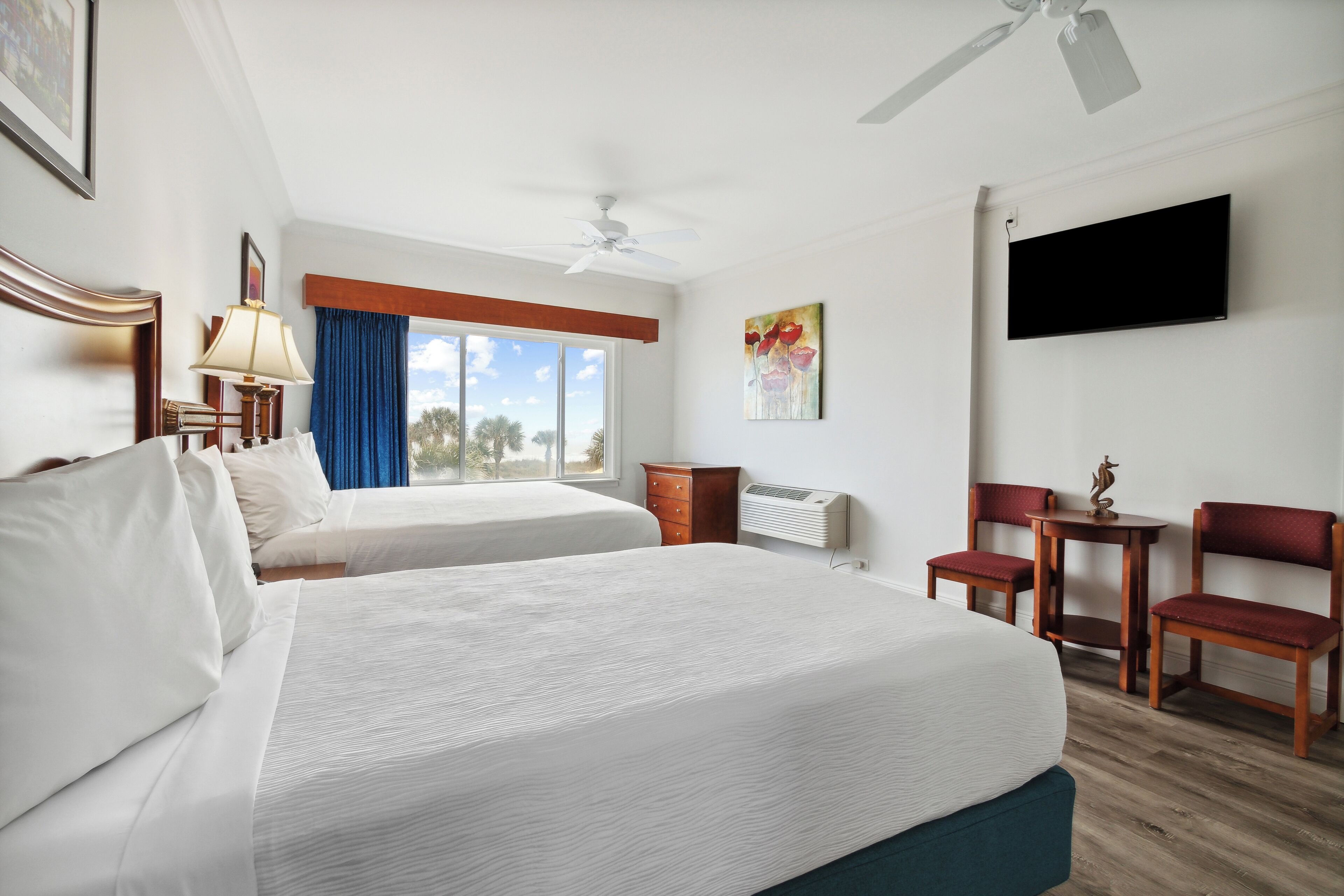 Habitación de hotel con dos camas, ventilador de techo, televisión de pantalla plana, escritorio con silla y ventana con cortinas azules.