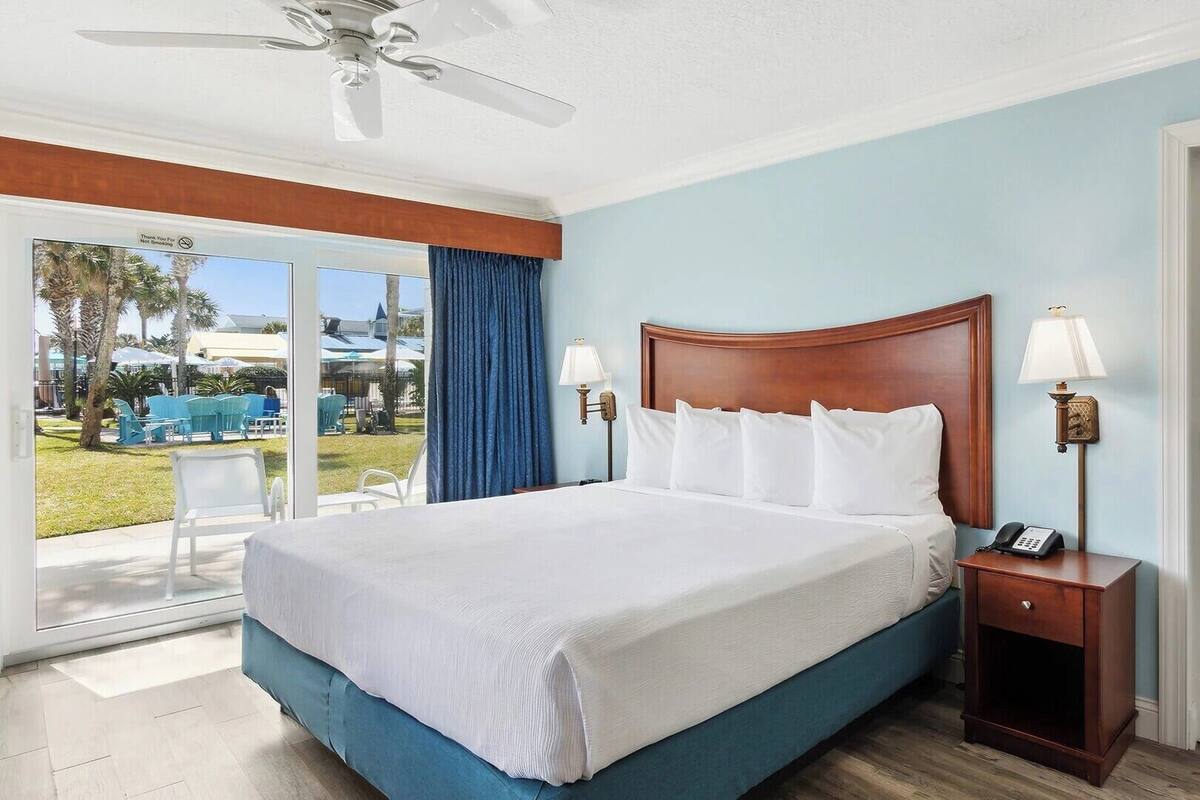A hotel room with a large bed, a nightstand with a lamp, and a view of the outdoor area.
