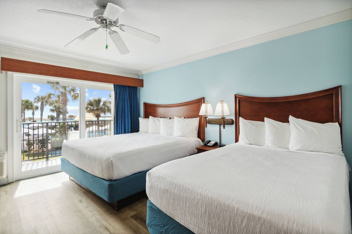 A hotel room with two beds, a ceiling fan, a nightstand with a lamp, and a view of palm trees through a sliding door.