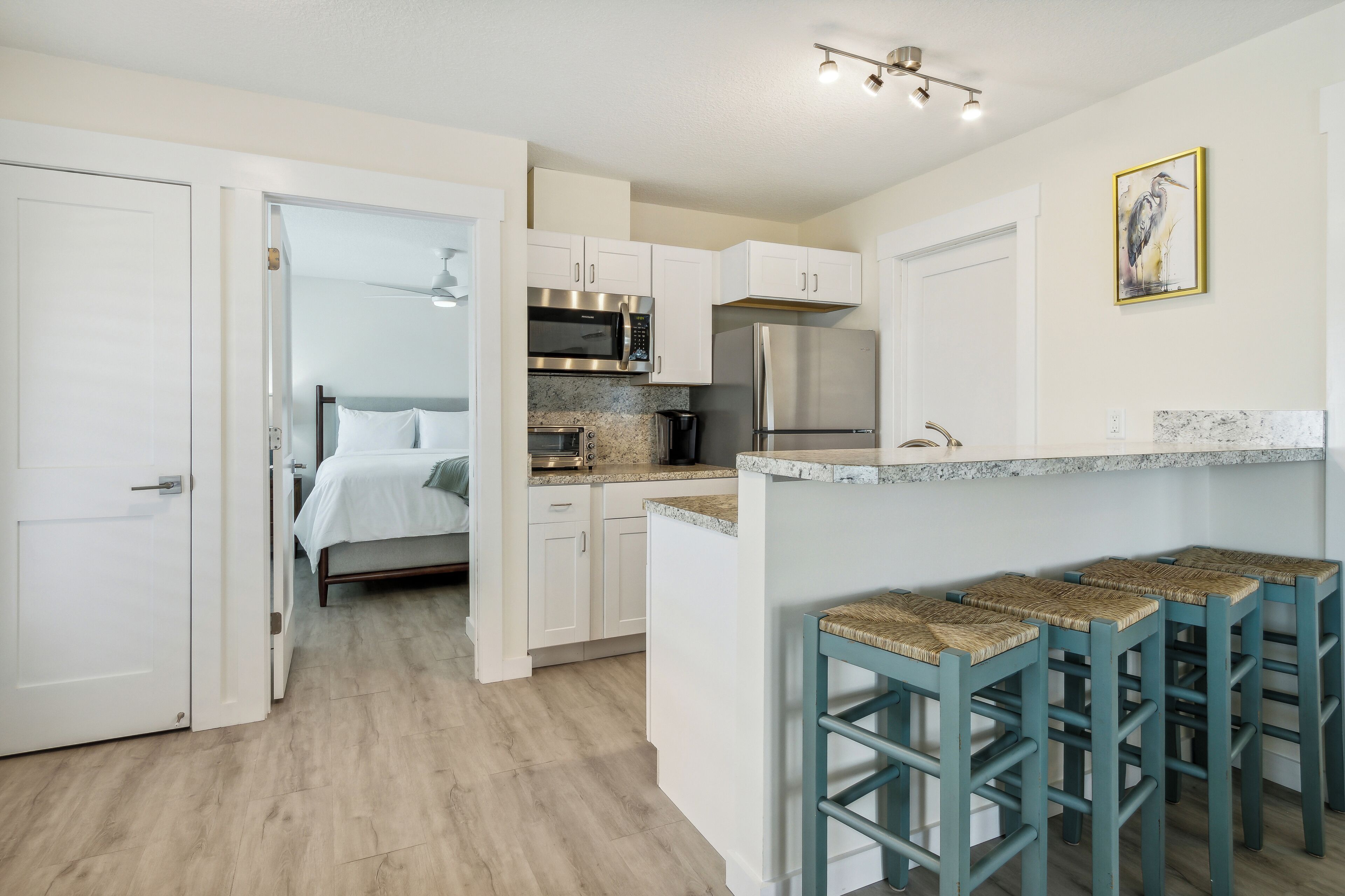 A compact kitchen with white cabinets, a stainless steel refrigerator, and a microwave. There is a breakfast bar with woven stools.