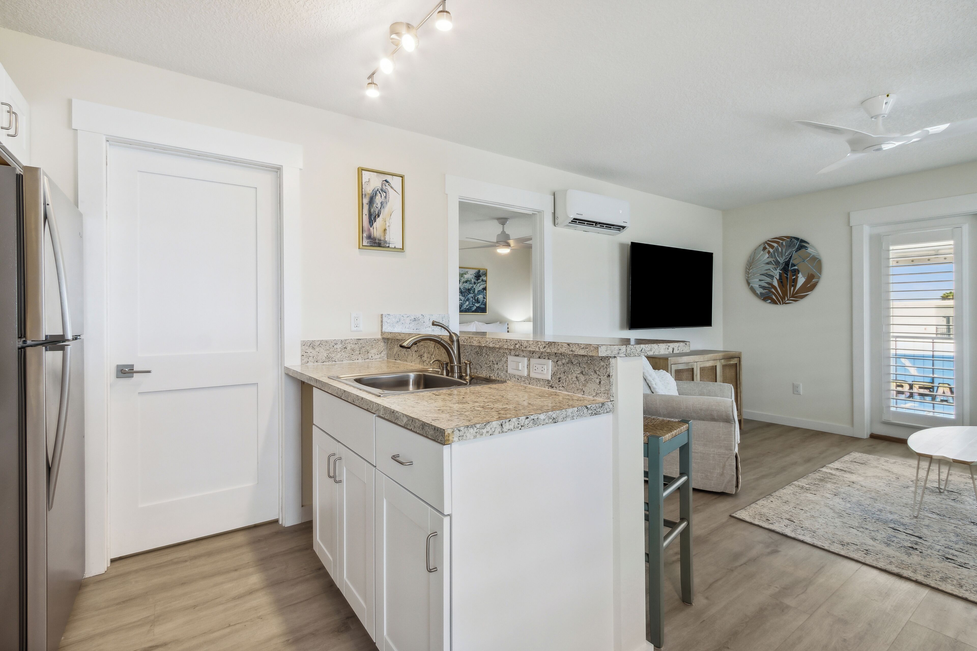 A modern kitchen with white cabinetry, granite countertops, and a built-in island. There is a dining area with a table and chairs, and a large flat-screen TV mounted on the wall.
