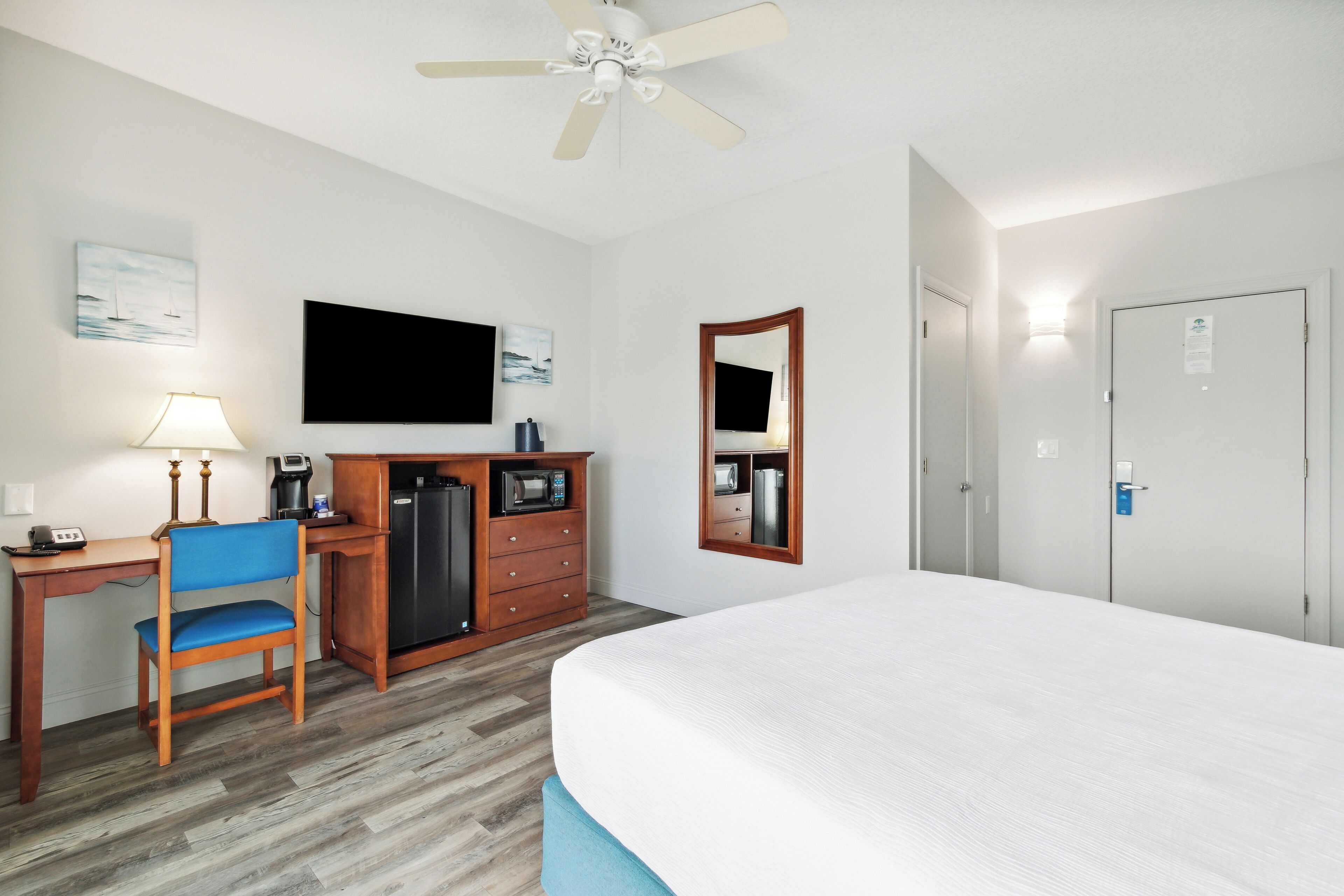 Een hotelkamer met een bed, bureau, stoel, kitchenette en een plafondventilator.