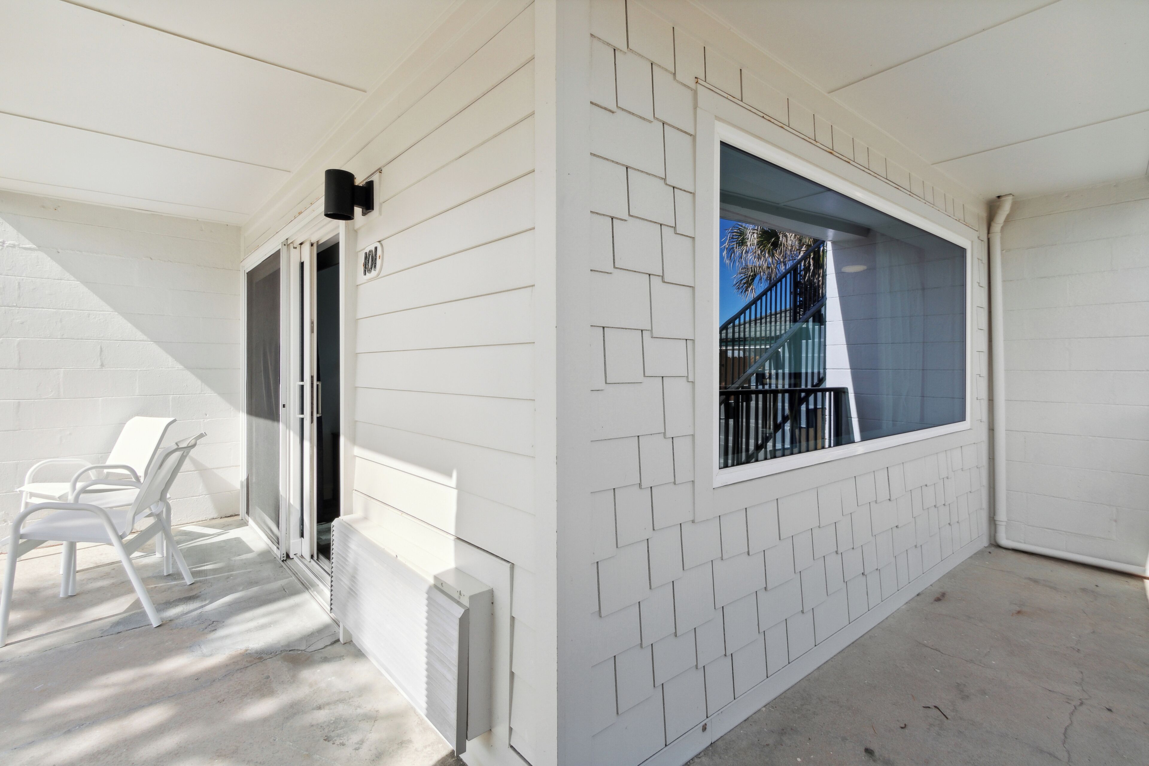 Une façade blanche avec une fenêtre et une porte, un sol en béton et deux chaises blanches.