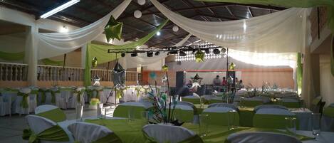 Un salón de banquetes con mesas dispuestas para un evento formal, cubierto con telas blancas y verdes y decoraciones en forma de estrella.