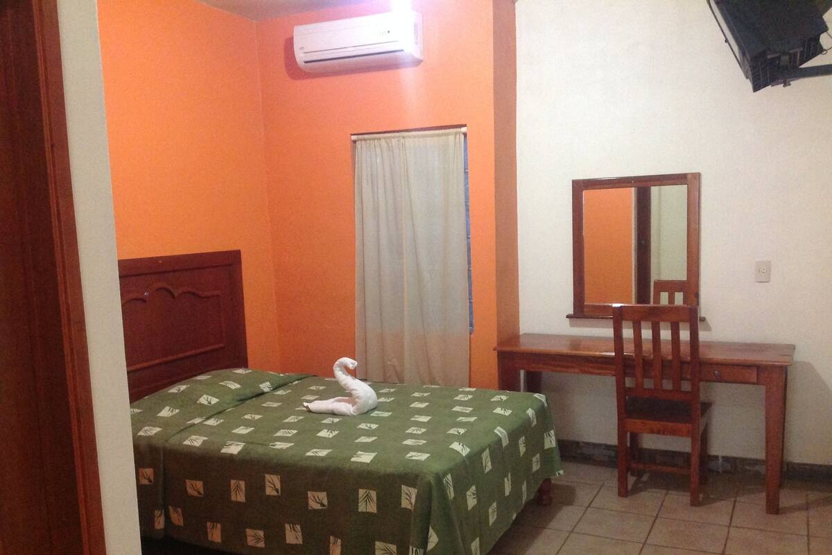 Una habitación con cama, escritorio, silla, ventana con cortinas y aire acondicionado.