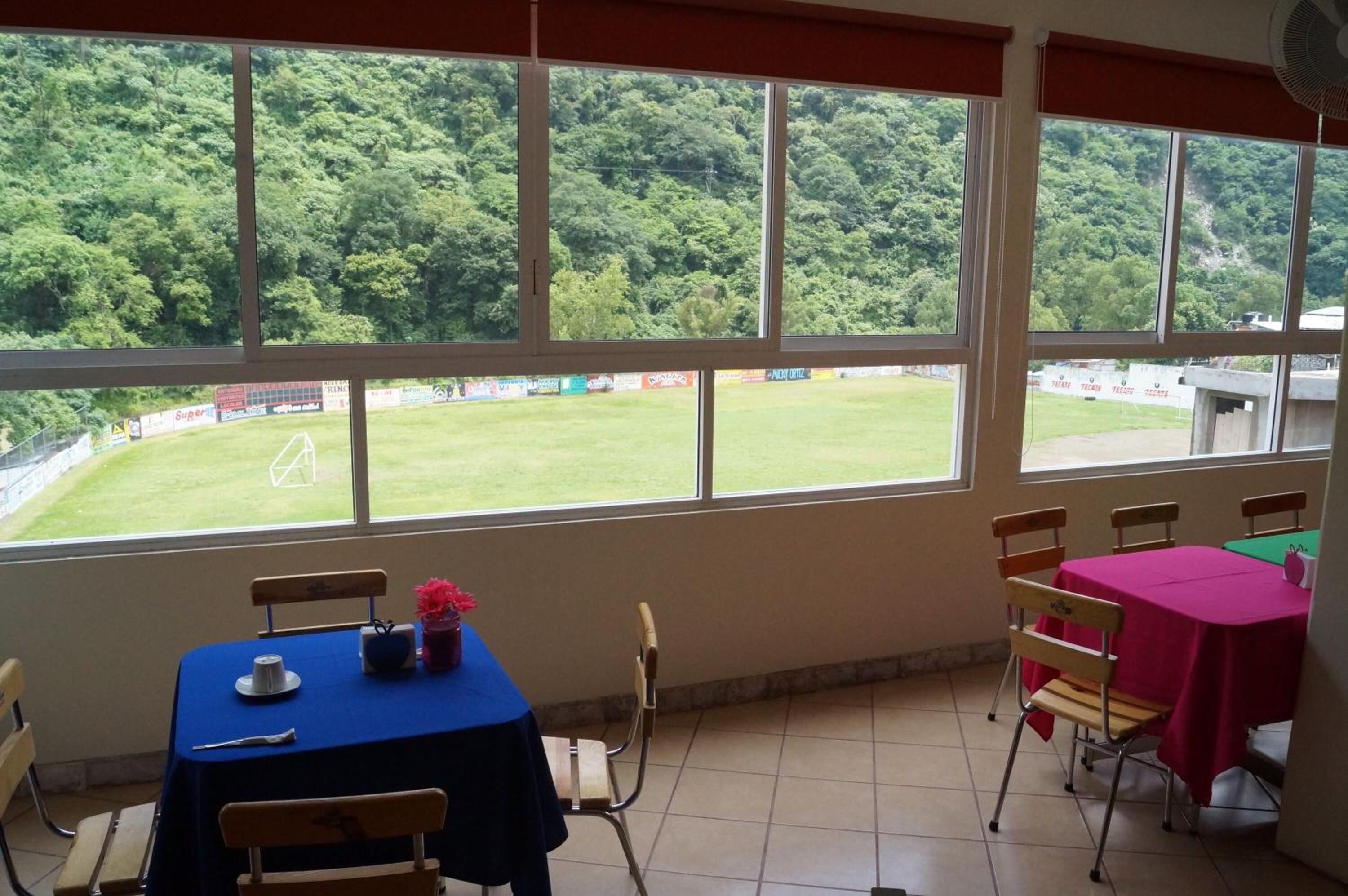 A room with large windows overlooking a grassy field, a table with a blue tablecloth, and wooden chairs.