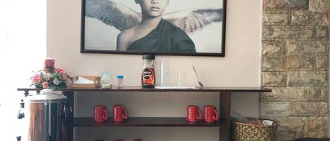 A living room with a television mounted on the wall, a coffee machine, and a shelf with red mugs.