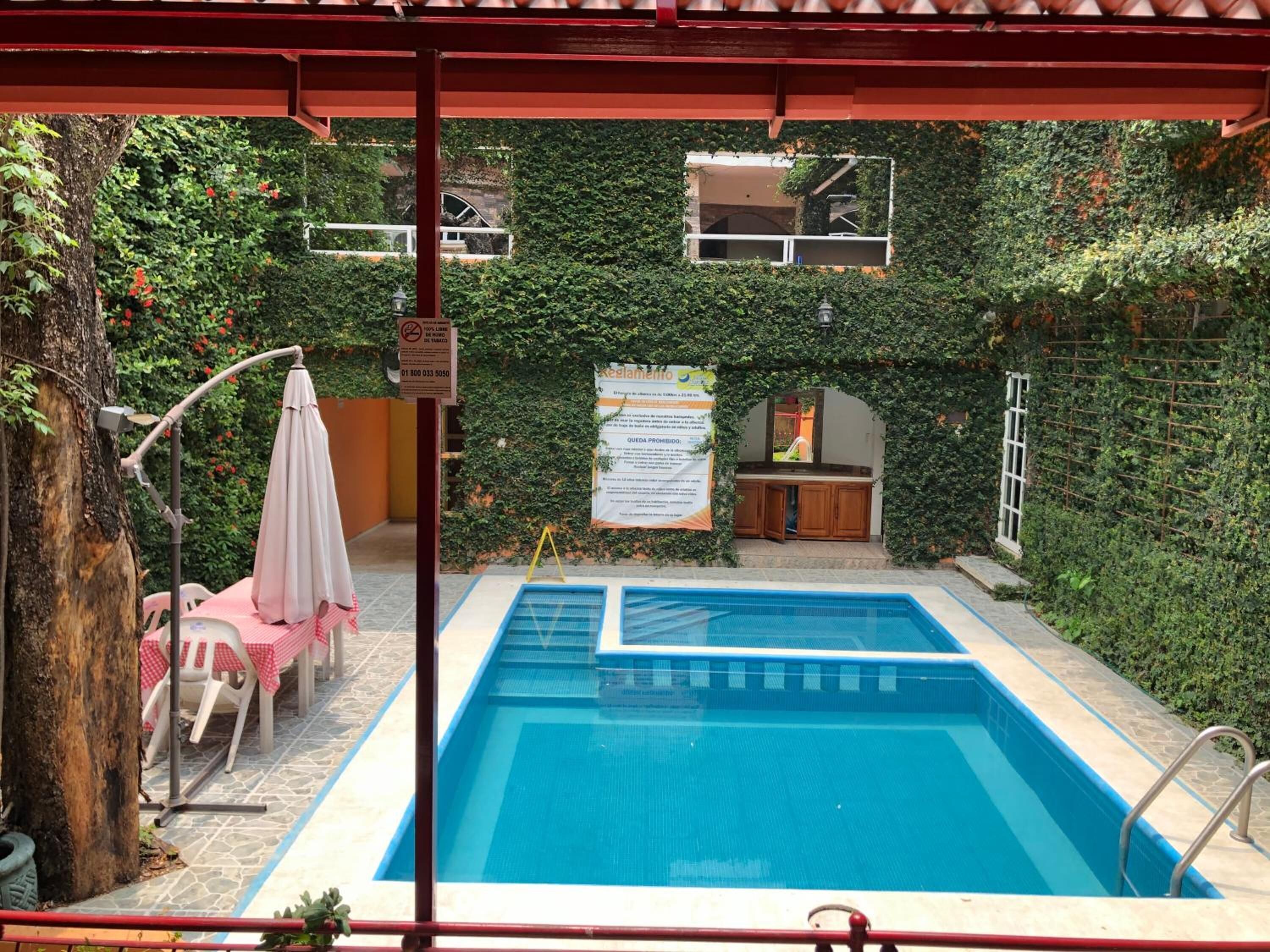 An outdoor swimming pool with a red pergola overhead, surrounded by lush greenery and a small wooden cabin.
