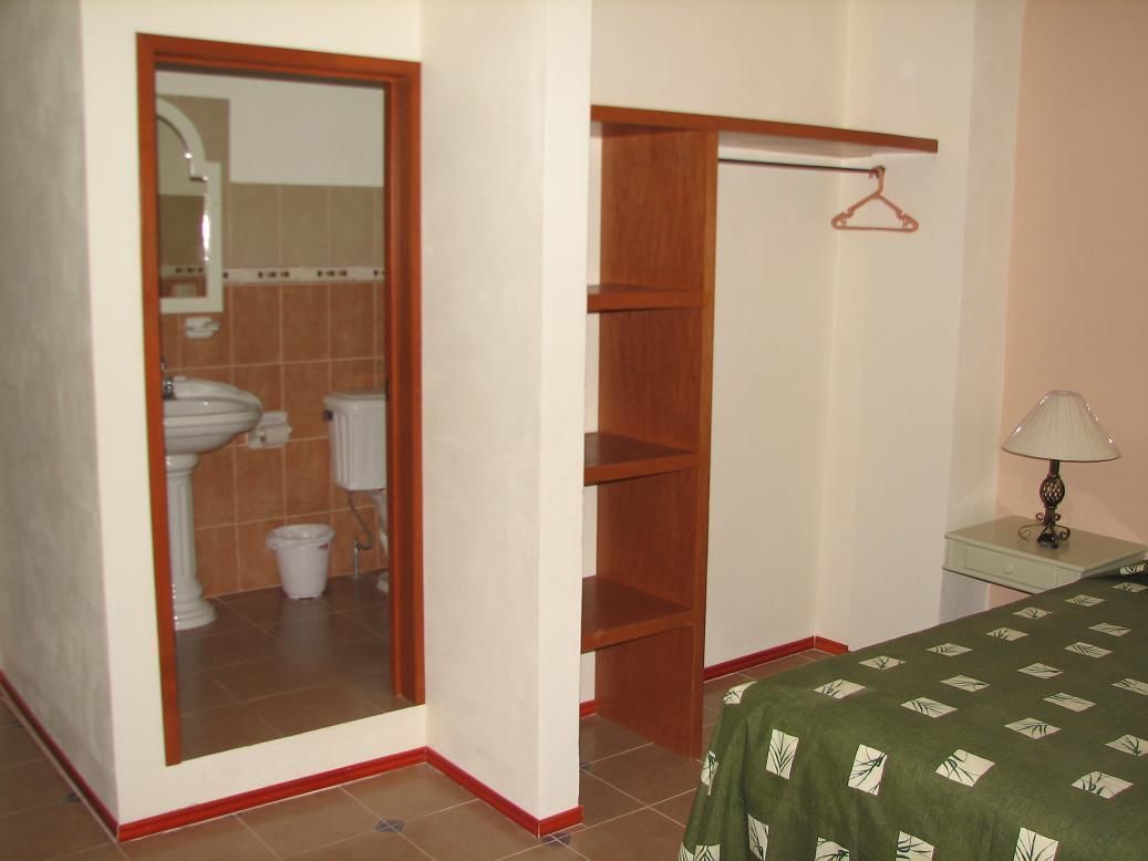 Une chambre à coucher comprenant un lit, une table de chevet, un miroir, une armoire en bois et une salle de bain visible à travers une porte ouverte.