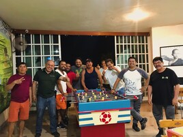 Eine Gruppe von Personen steht um einen Fußballtisch in einem Raum mit großen Fenstern und einem Wandventilator.