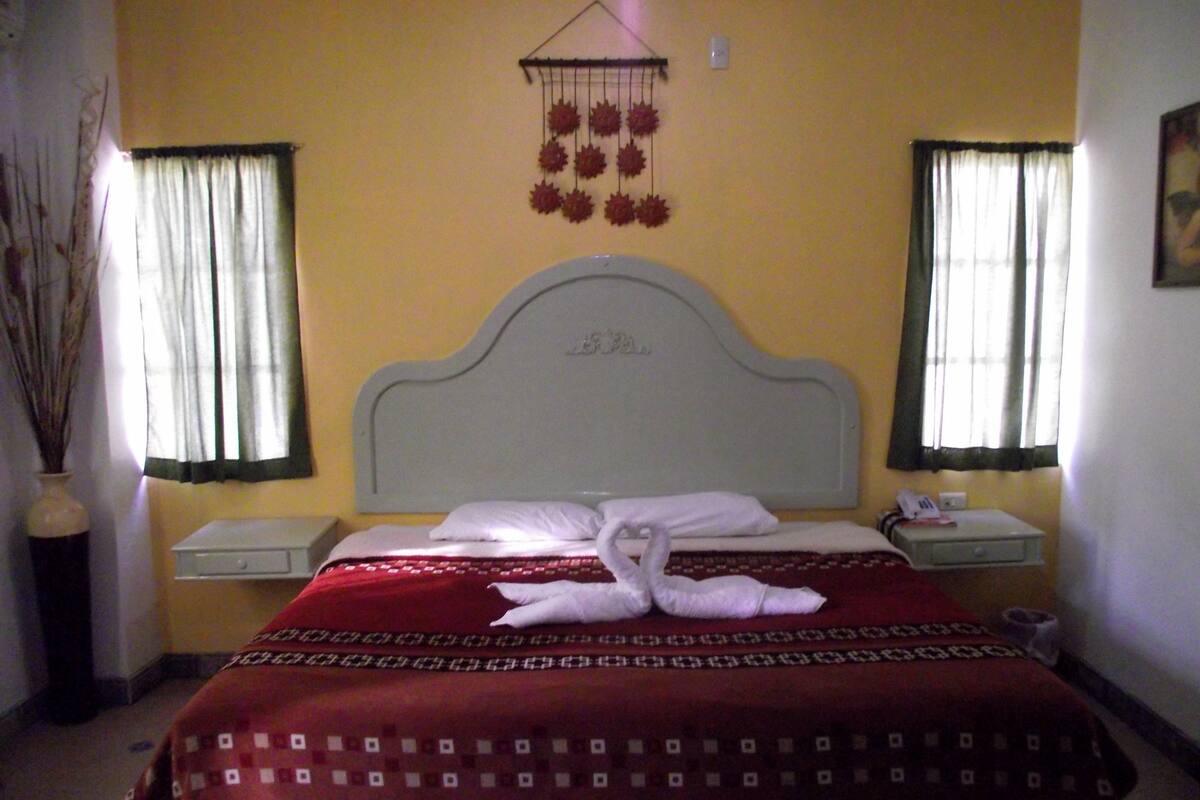 Una habitación de hotel con cama, dos mesitas de noche, un adorno en la pared y una ventana con cortinas.