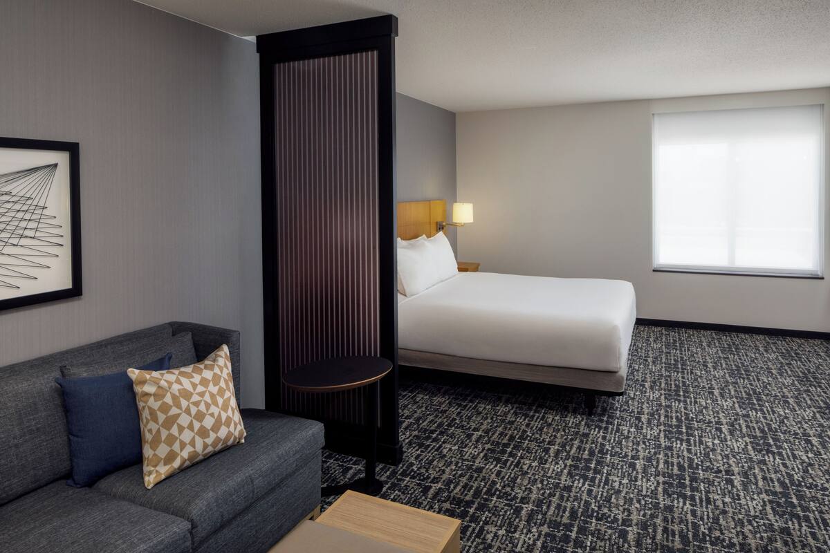 A hotel room with a grey sofa, a bed with white linens, a small round table, and a large window with blinds.
