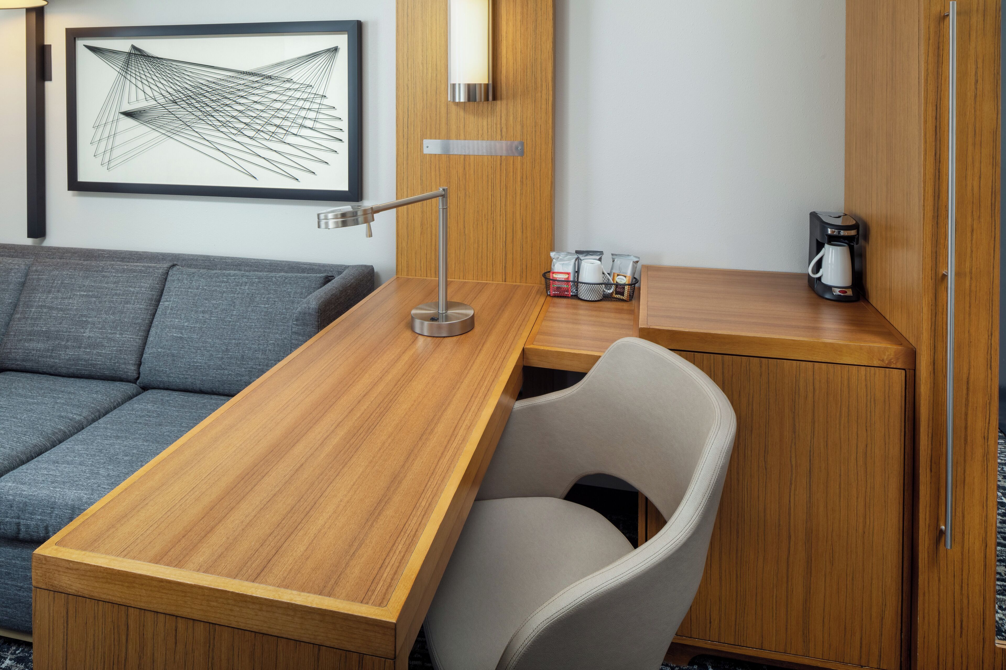 Una habitación de hotel moderna con un escritorio de madera, un sofá gris, una silla blanca, una cafetera y una obra de arte abstracta enmarcada en la pared.