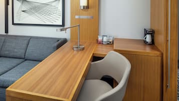 Una habitación de hotel moderna con un escritorio de madera, un sofá gris, una silla blanca, una cafetera y una obra de arte abstracta enmarcada en la pared.