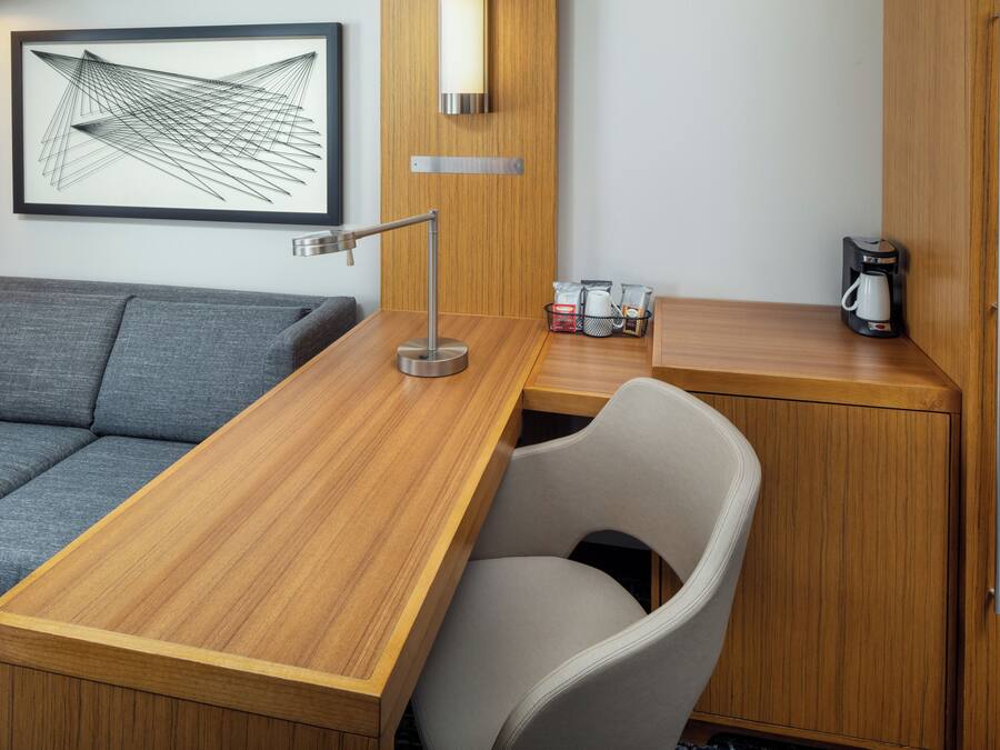 Una habitación de hotel moderna con un escritorio de madera, un sofá gris, una silla blanca, una cafetera y una obra de arte abstracta enmarcada en la pared.