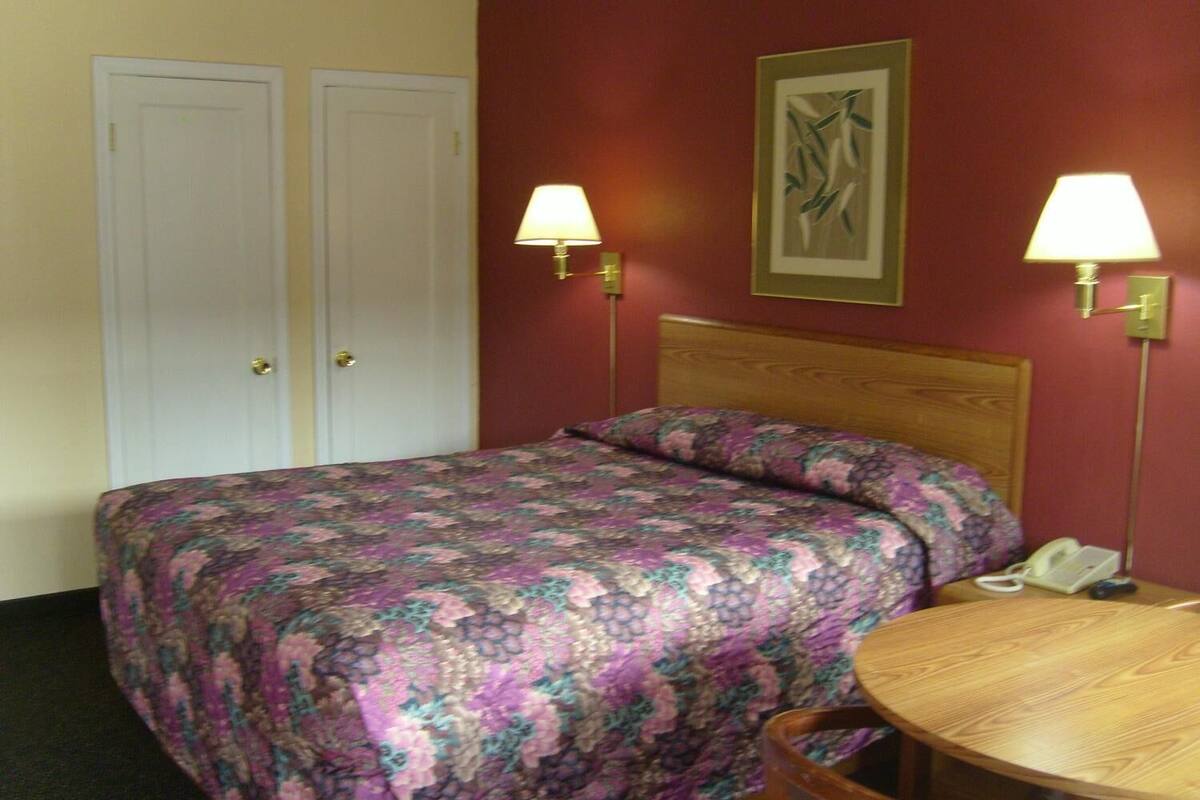 A hotel room with a bed, two bedside lamps, a wooden headboard, a round wooden table, and a telephone on the table.