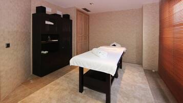 A room with a bed, a dark wooden wardrobe, a small table with a towel, and a window with blinds.