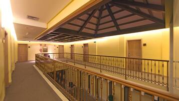 A hotel corridor with a wooden railing, a high ceiling with a truss structure, and doors leading to rooms.