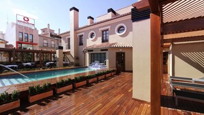 A wooden deck with a pool, outdoor seating, and a building with a Vodafone sign. - Hotel Casa Consistorial (Fuengirola)