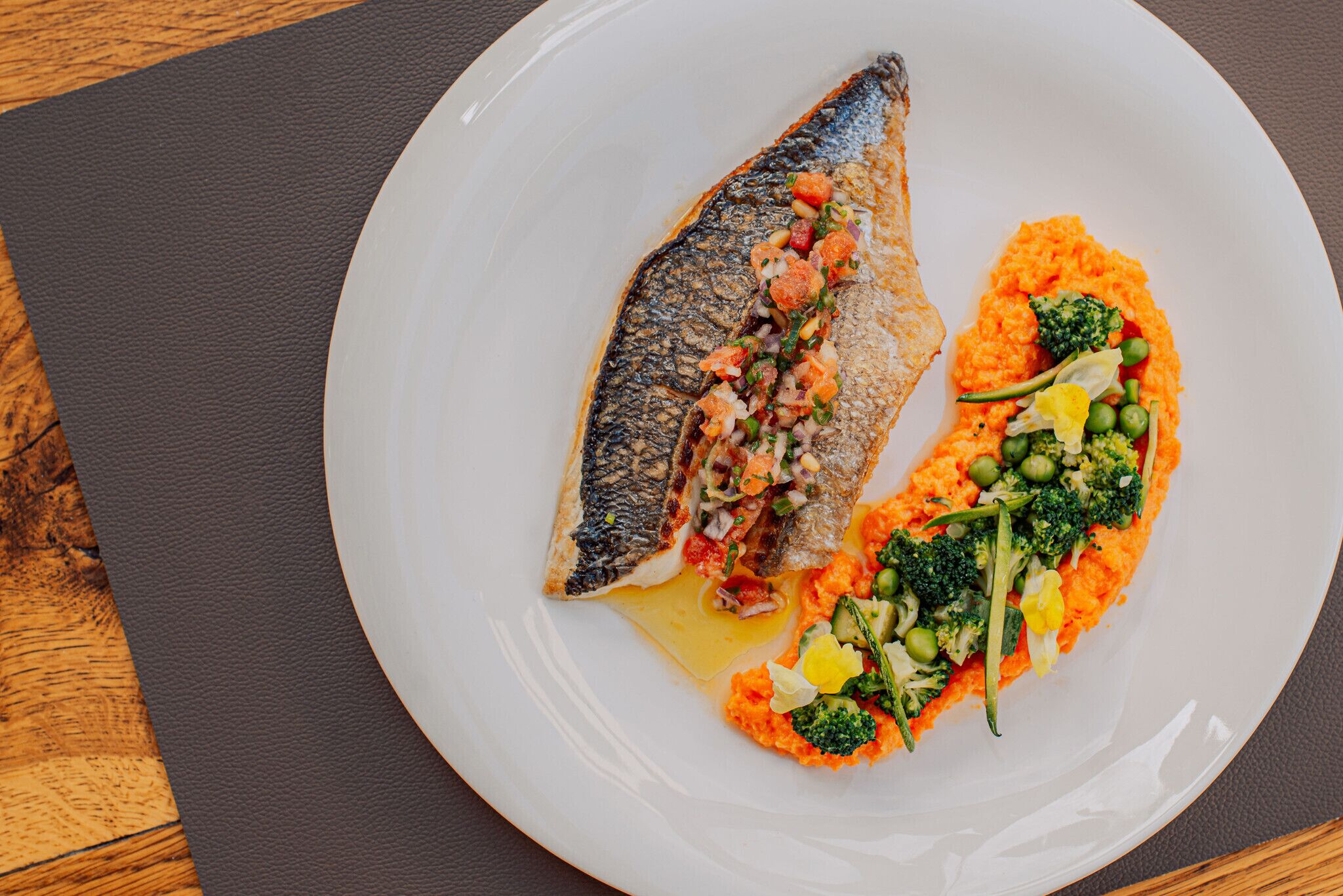 A white plate with a fish and vegetables on a dark mat.
