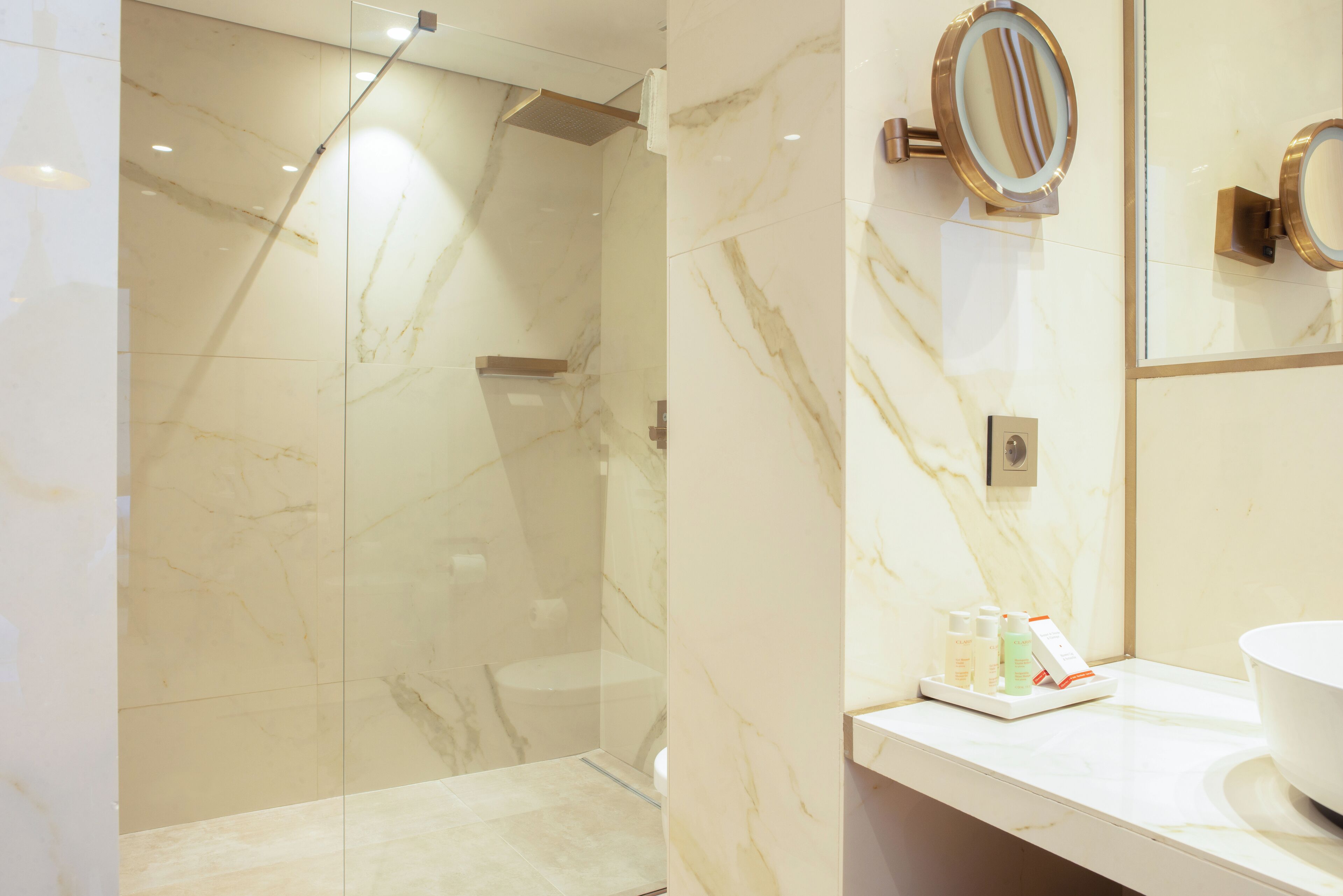 Un baño moderno con ducha envuelta en vidrio, paredes de mármol y un tocador con artículos de aseo.