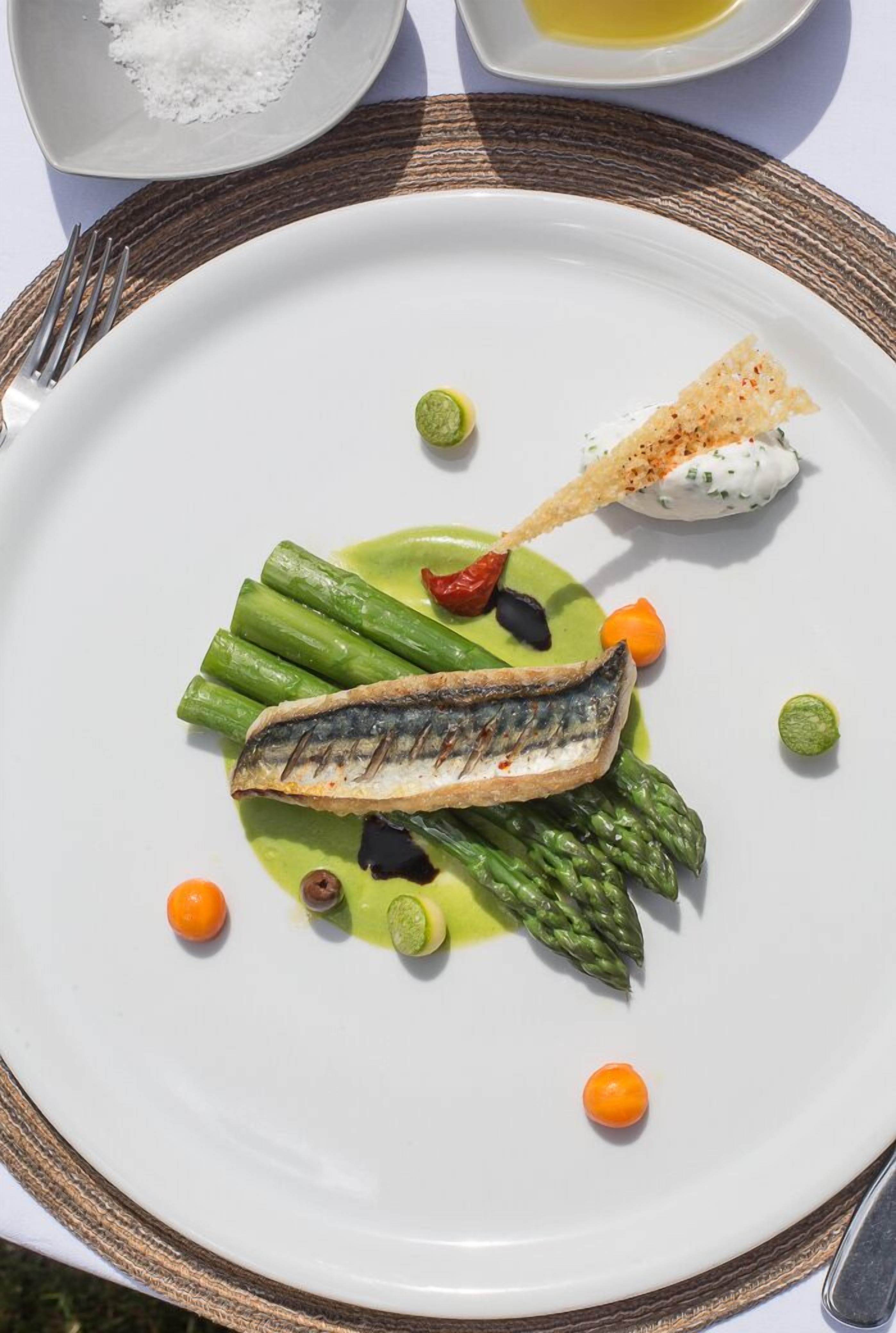 En vit tallrik med en grillad fisk, sparris och en grön sås, garnerad med små orange och gröna dekorationer.