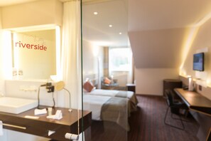 A hotel room with a bed, a desk, a chair, a TV, and a bathroom with a mirror and sink.