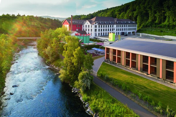 A riverside hotel with a modern building, a bridge, and lush greenery.