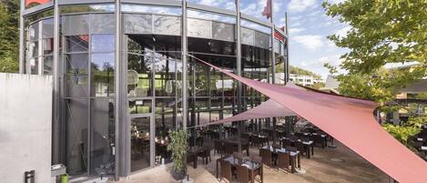 A modern building with a glass facade, outdoor seating area, and red flags on top.