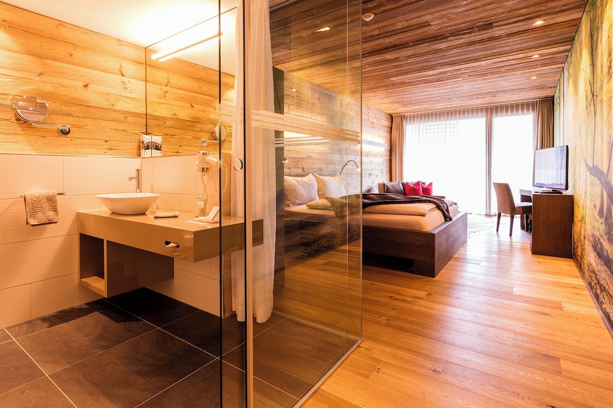 Habitación de hotel moderna con interior de madera, cabina de ducha de vidrio, cama con televisor y escritorio con computadora.