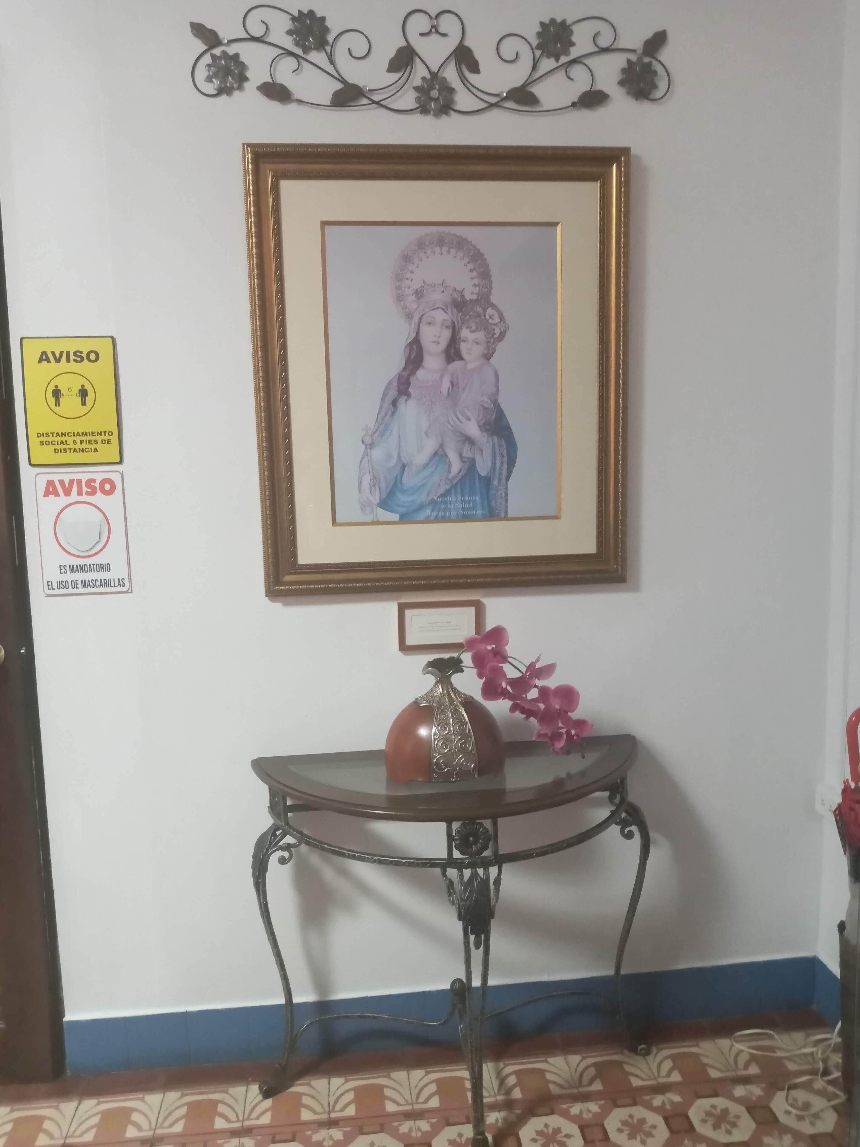 Un cuadro religioso enmarcado en una pared, una pequeña mesa con un florero y flores, y dos carteles amarillos con recomendaciones sobre distanciamiento social y el uso de mascarillas.