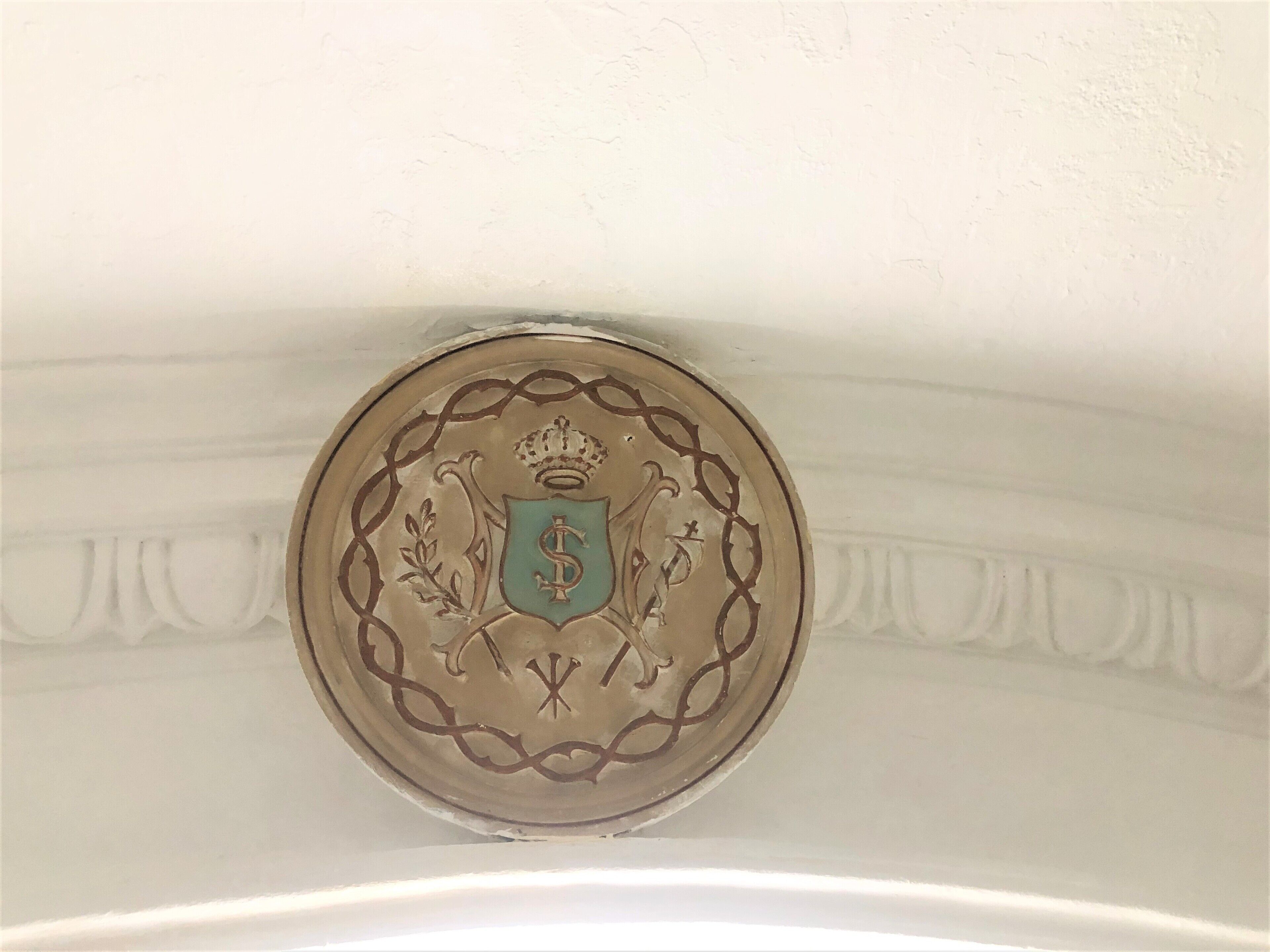 A decorative emblem with a crown, shield, and fleur-de-lis on a white wall.