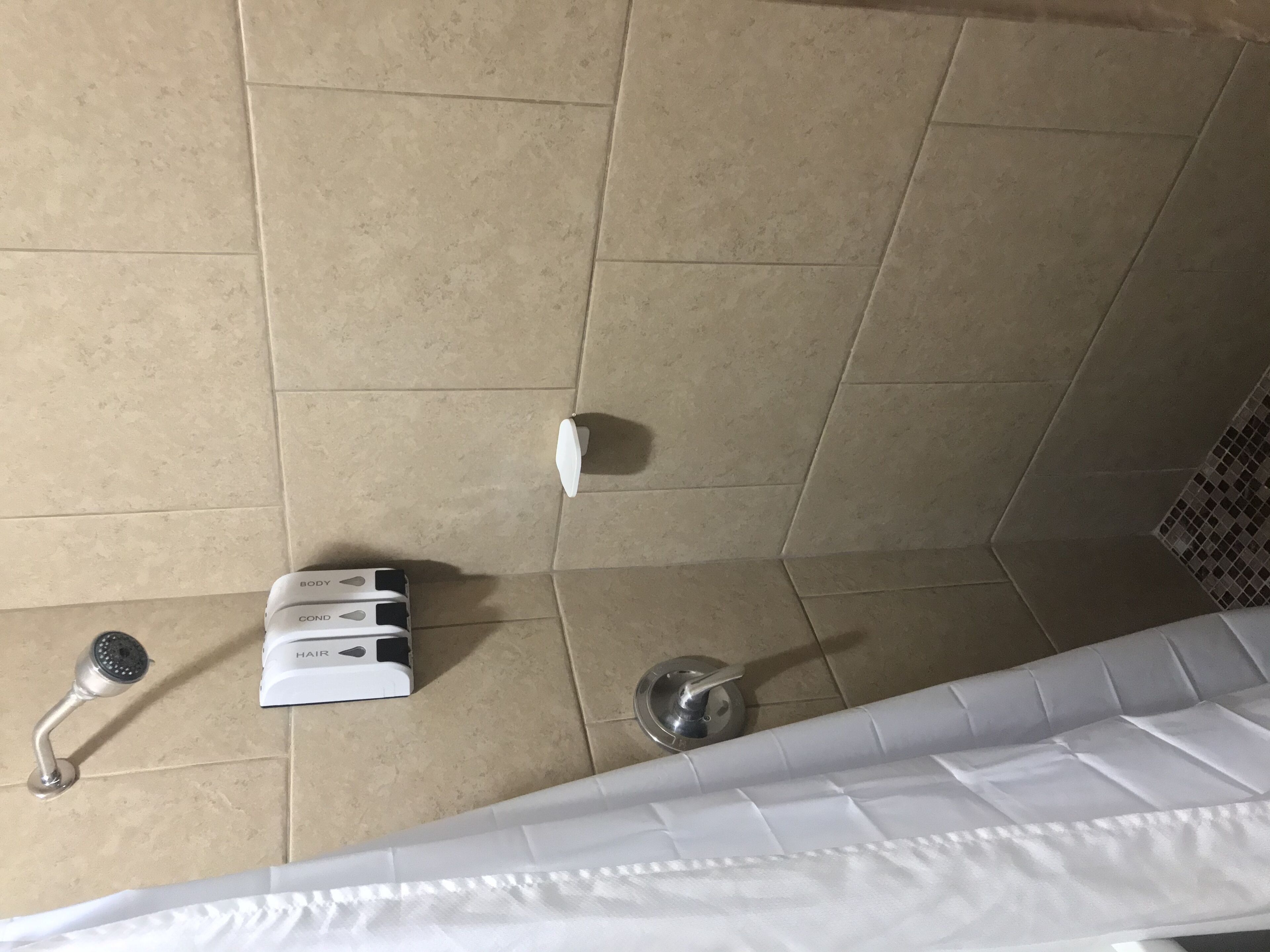 A bathroom with beige tiles, a white shower curtain, a handheld showerhead, a handheld showerhead, and a white box with the text "HARD" and "COND".