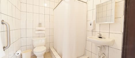 Un baño con azulejos blancos, un inodoro, una cortina de ducha, un lavabo y un pasamanos.