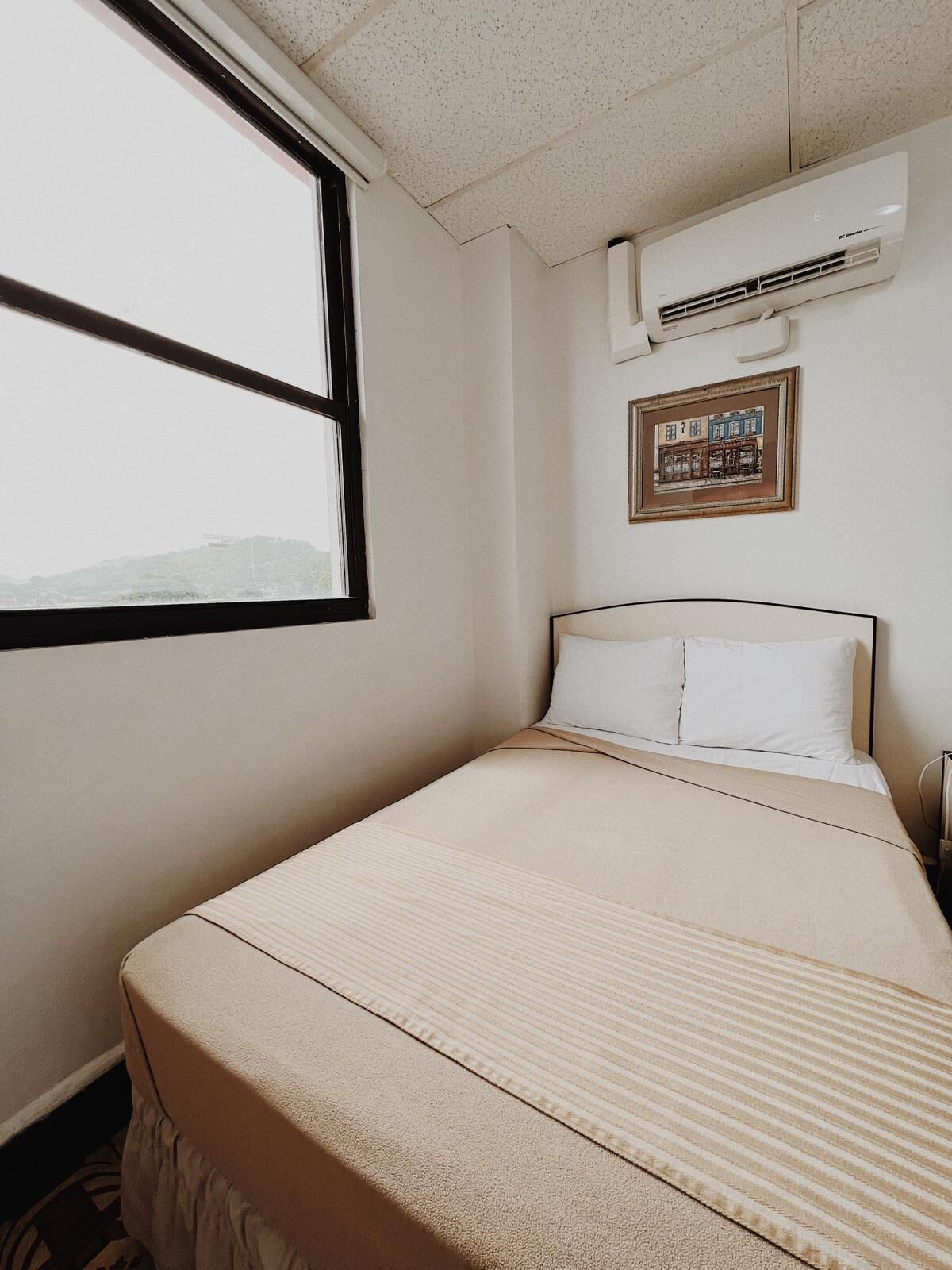 Une chambre d’hôtel avec un lit, une fenêtre, un appareil de climatisation et un tableau encadré au mur.
