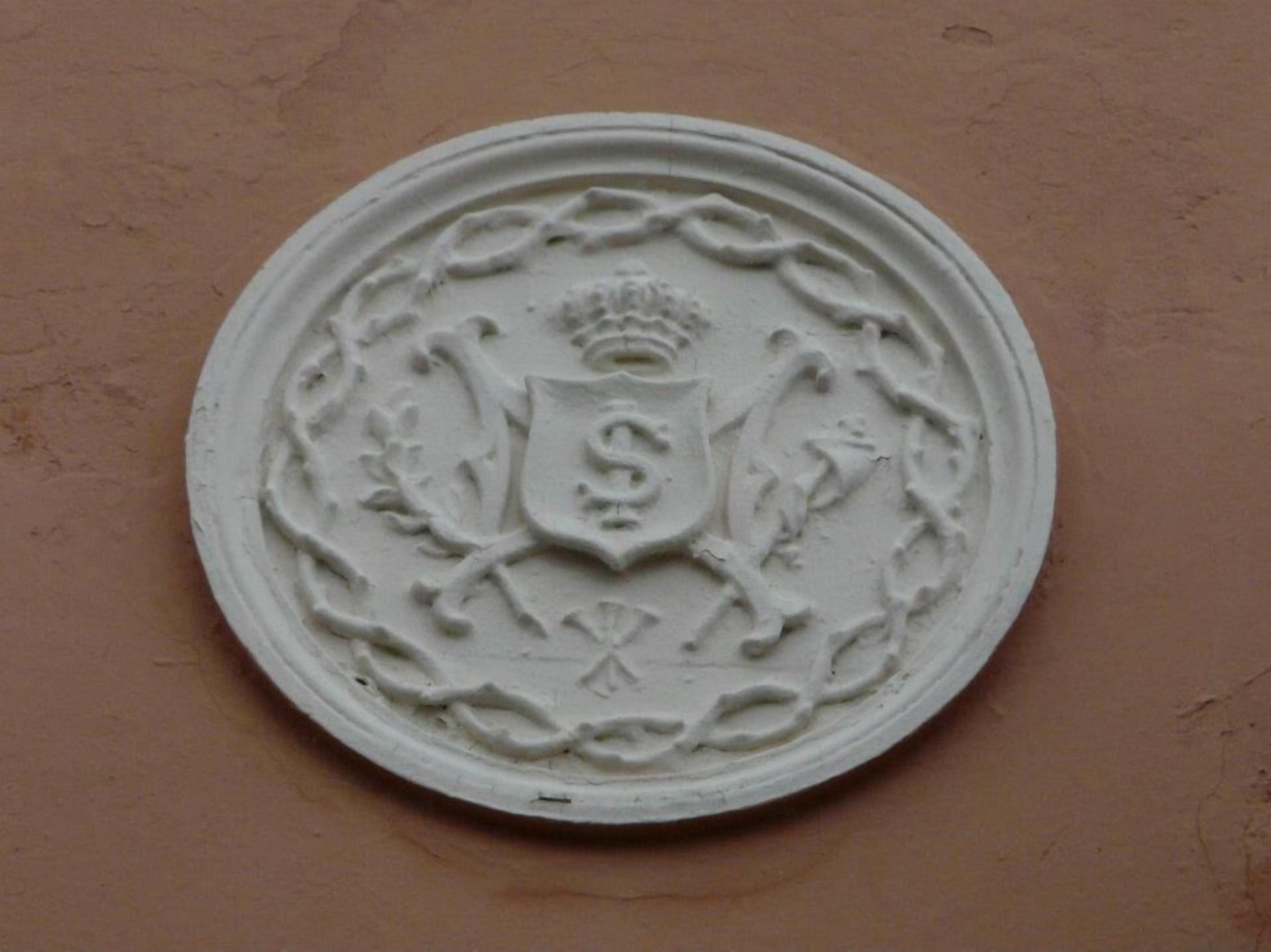 Ein kreisförmiges Emblem mit Schild und Krone vor einer schlichten Wand.