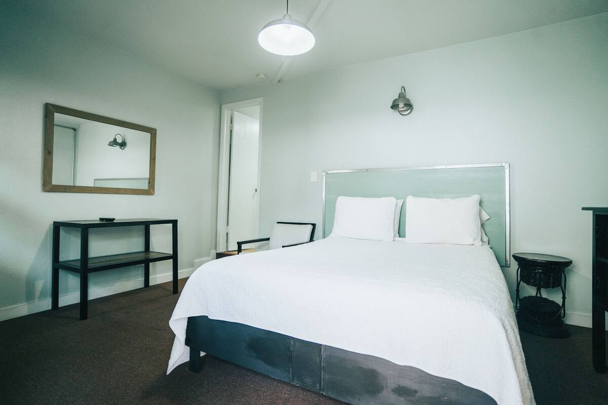 Ein Schlafzimmer mit einem Bett, einem Nachttisch, einem Sessel, einem kleinen Tisch, einem Spiegel und einer Lampe.