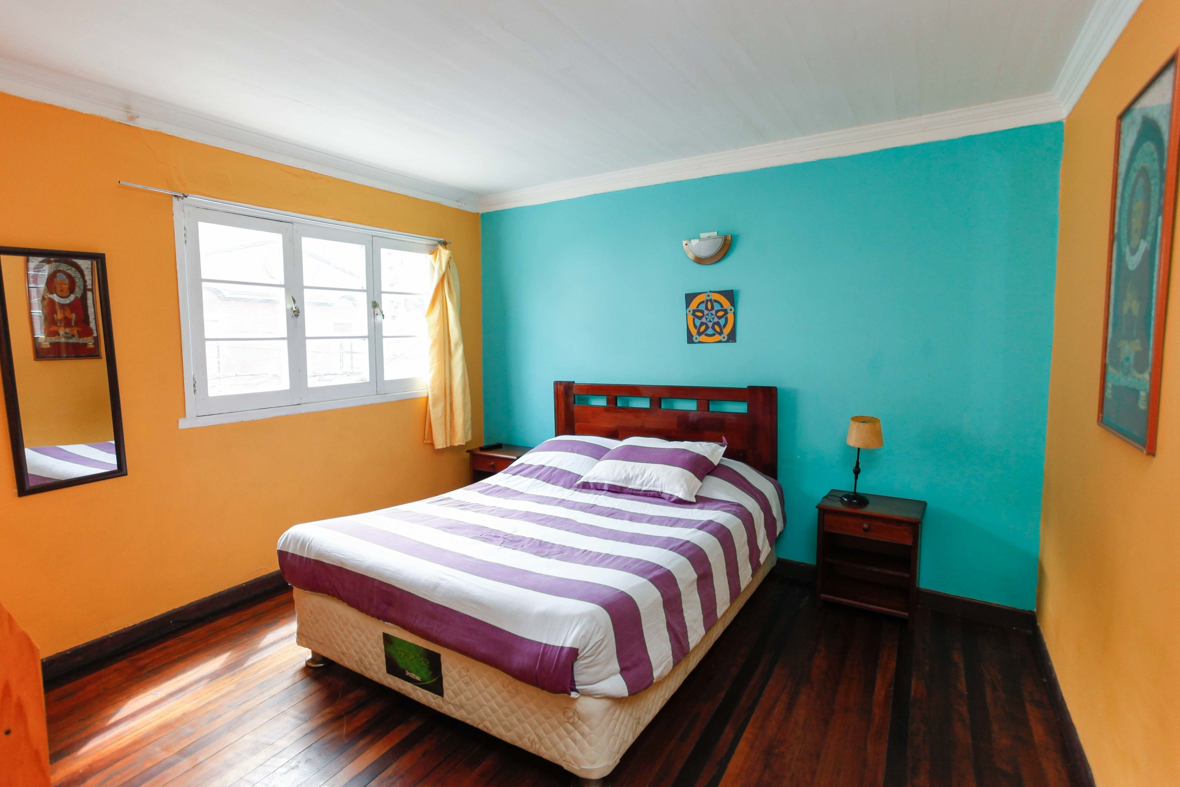 A bedroom with a bed, a nightstand, a lamp, a window with curtains, and a wall with a painting.