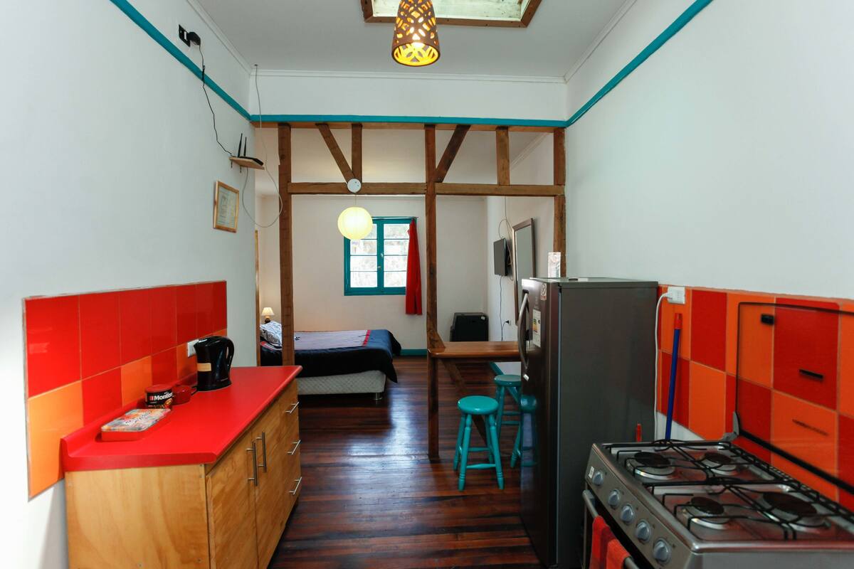 A compact kitchen with a stove, a sink, and a countertop. There is a bed with a red blanket and a window with curtains.