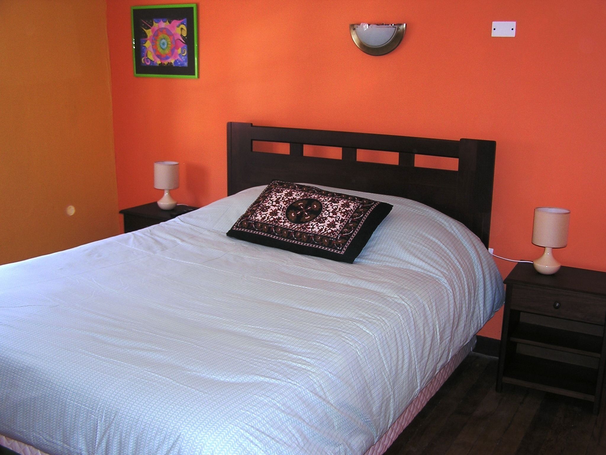 Una cama con cubrecama blanco y un cojín decorativo, dos mesitas de noche con lámparas, y una pared con un cuadro colorido y una lámpara de pared.