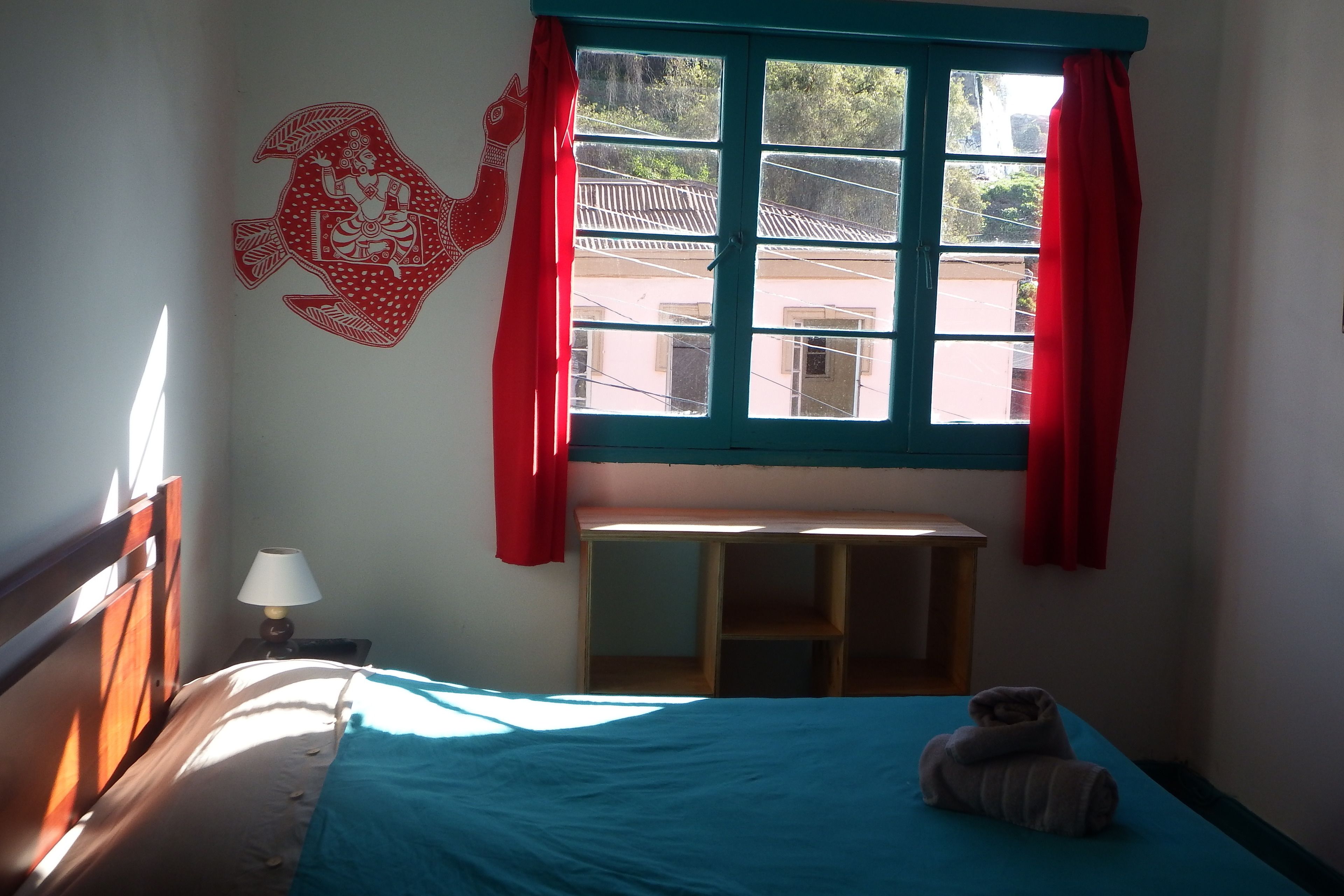 Um quarto com uma cama, uma janela com cortinas vermelhas, um adorno na parede e uma mesinha.
