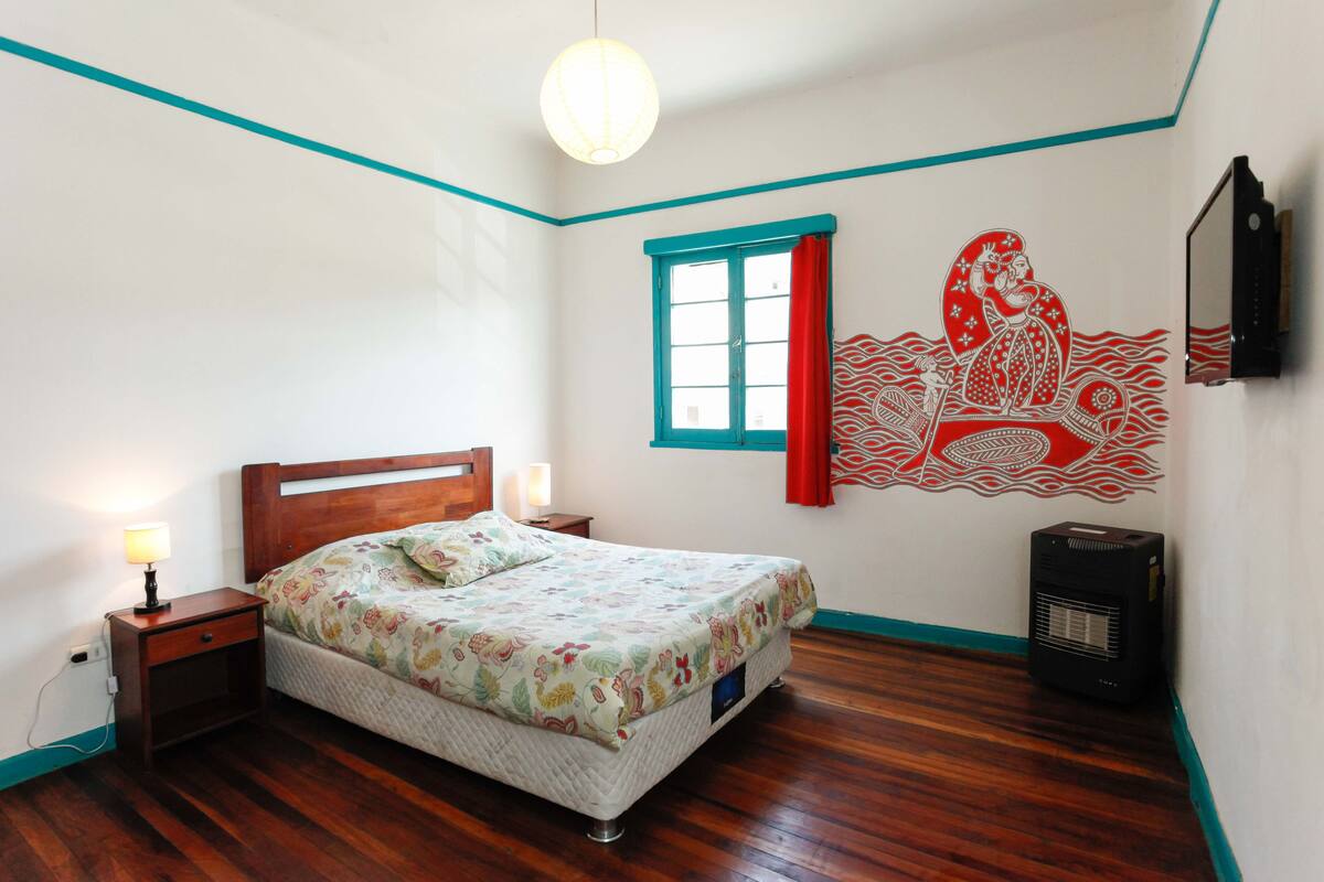 A bedroom with a bed, a nightstand, a wall-mounted heater, and a window with red curtains.
