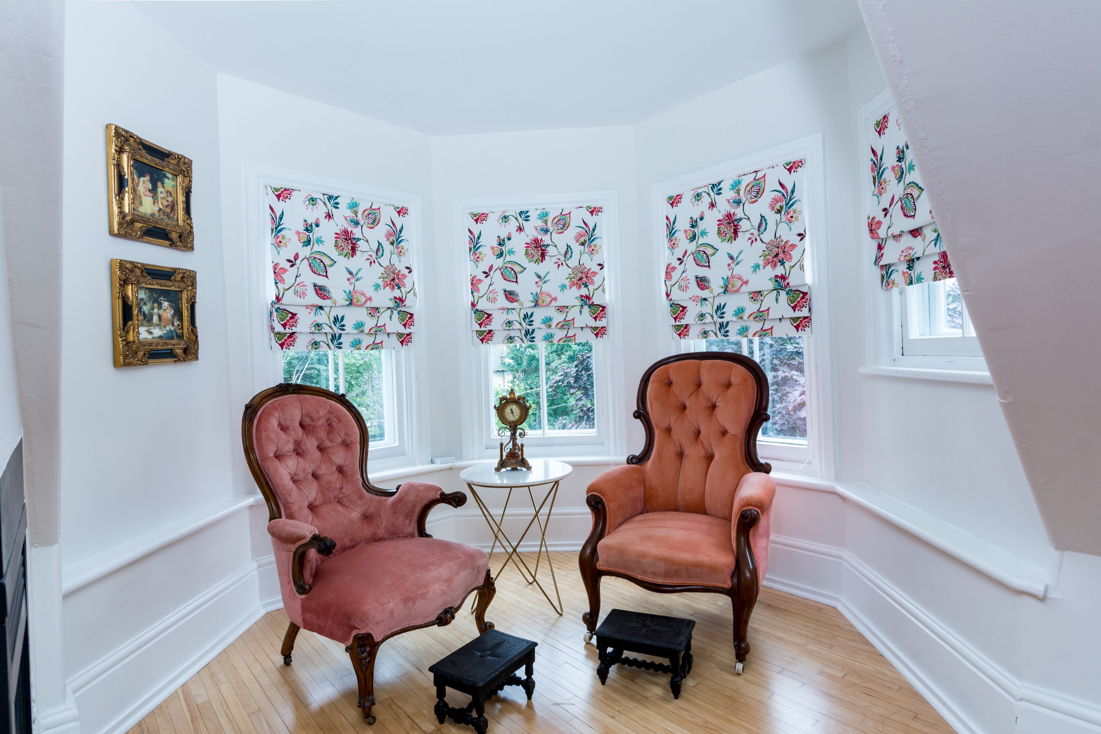 A room with two antique chairs, a small table with a clock, and floral-patterned window valances.