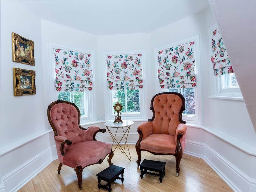 A room with two antique chairs, a small table with a clock, and floral-patterned window valances.