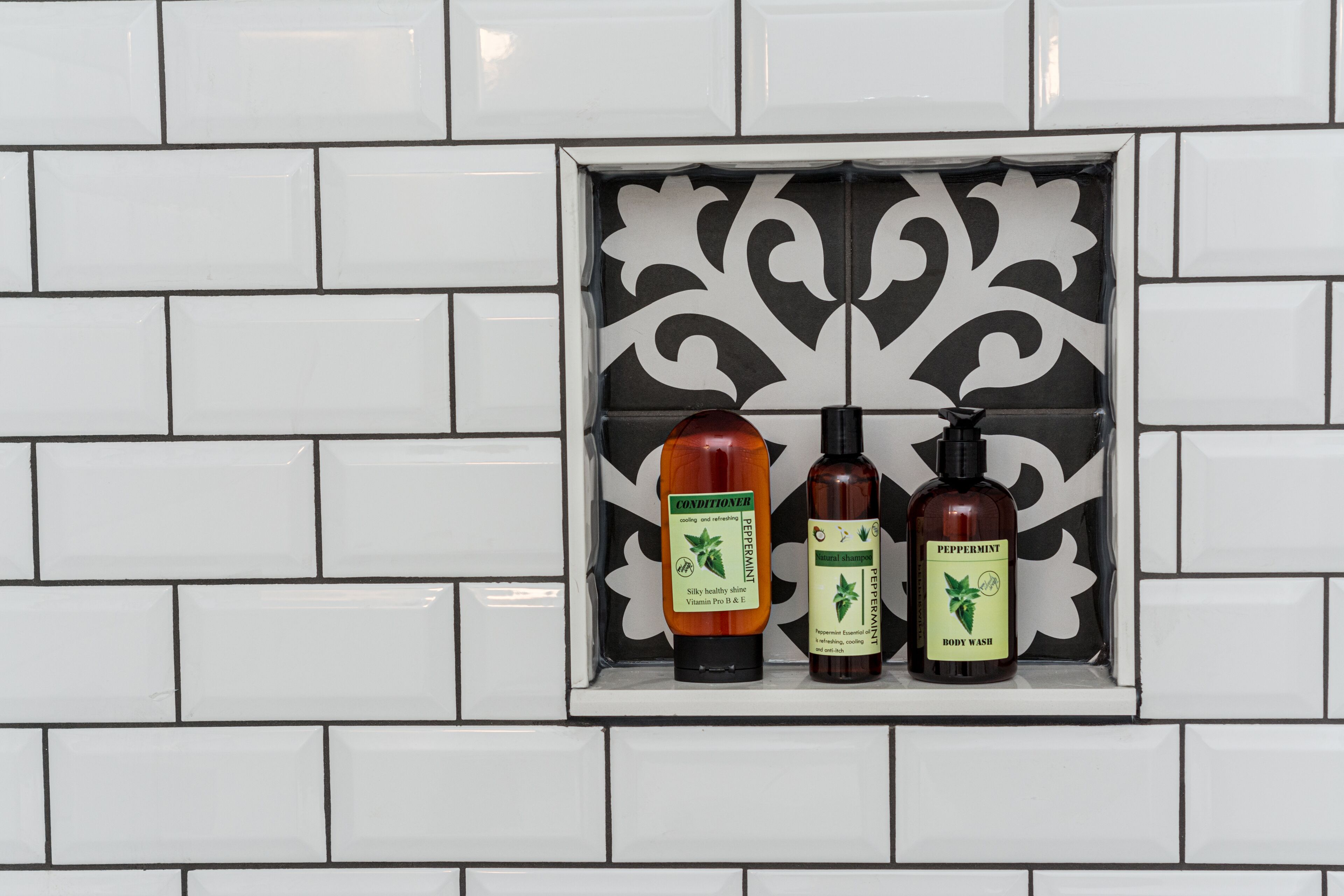A bathroom with white tiles and a decorative black and white tile accent piece. Three bottles of body wash are displayed on a shelf.