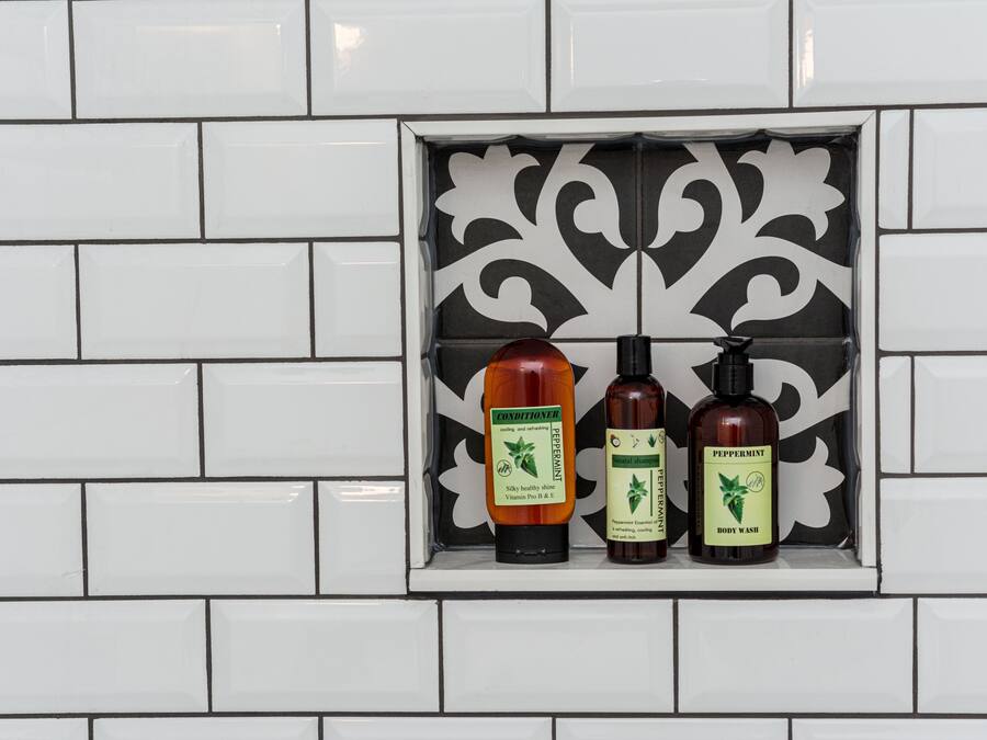 Baño con azulejos blancos y un detalle decorativo de baldosas blanco y negro. Se exhiben tres botellas de jabón corporal en una repisa.