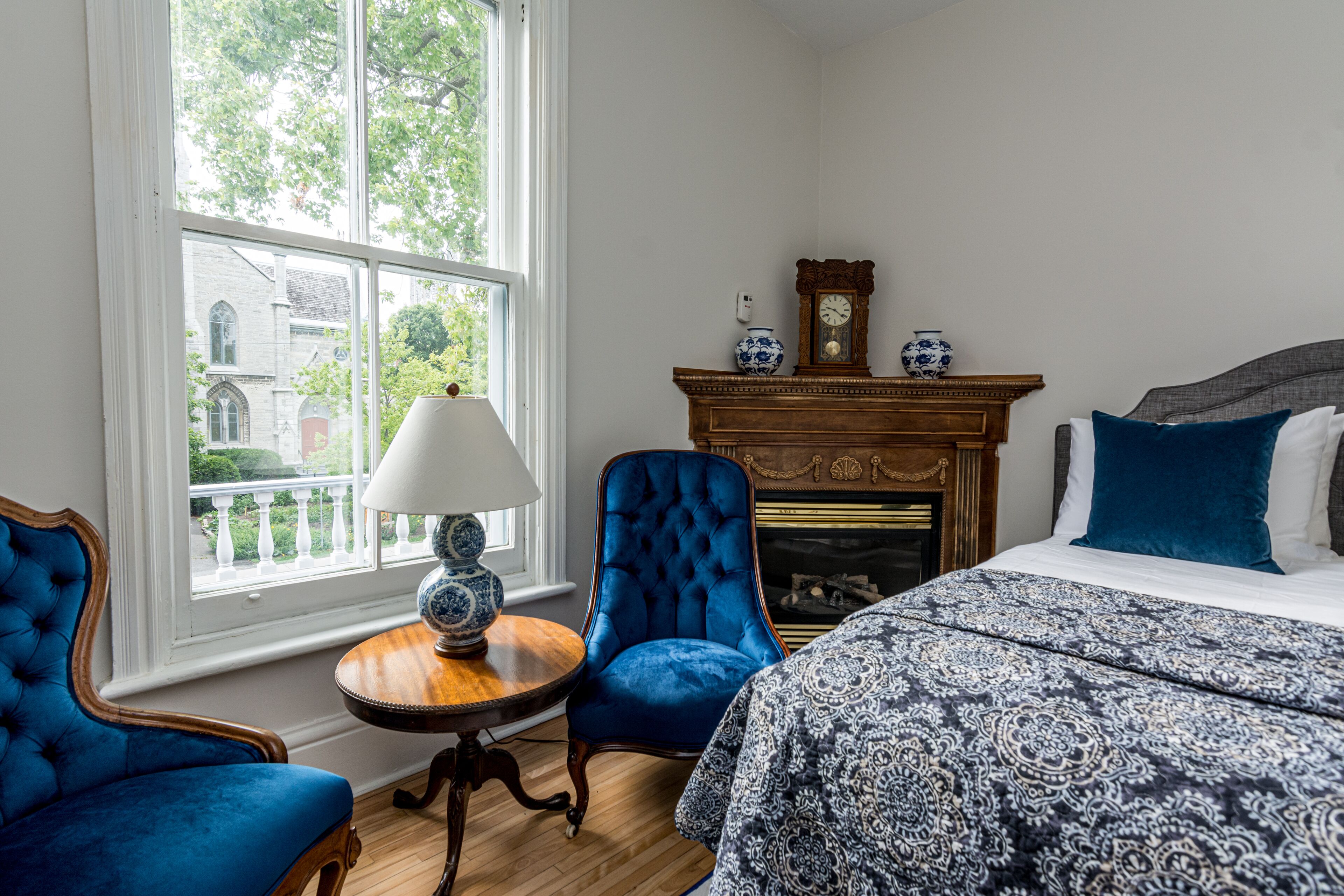 Una habitación con un gran ventanal, una chimenea, una cama con una colcha estampada y dos sillones tapizados en azul.